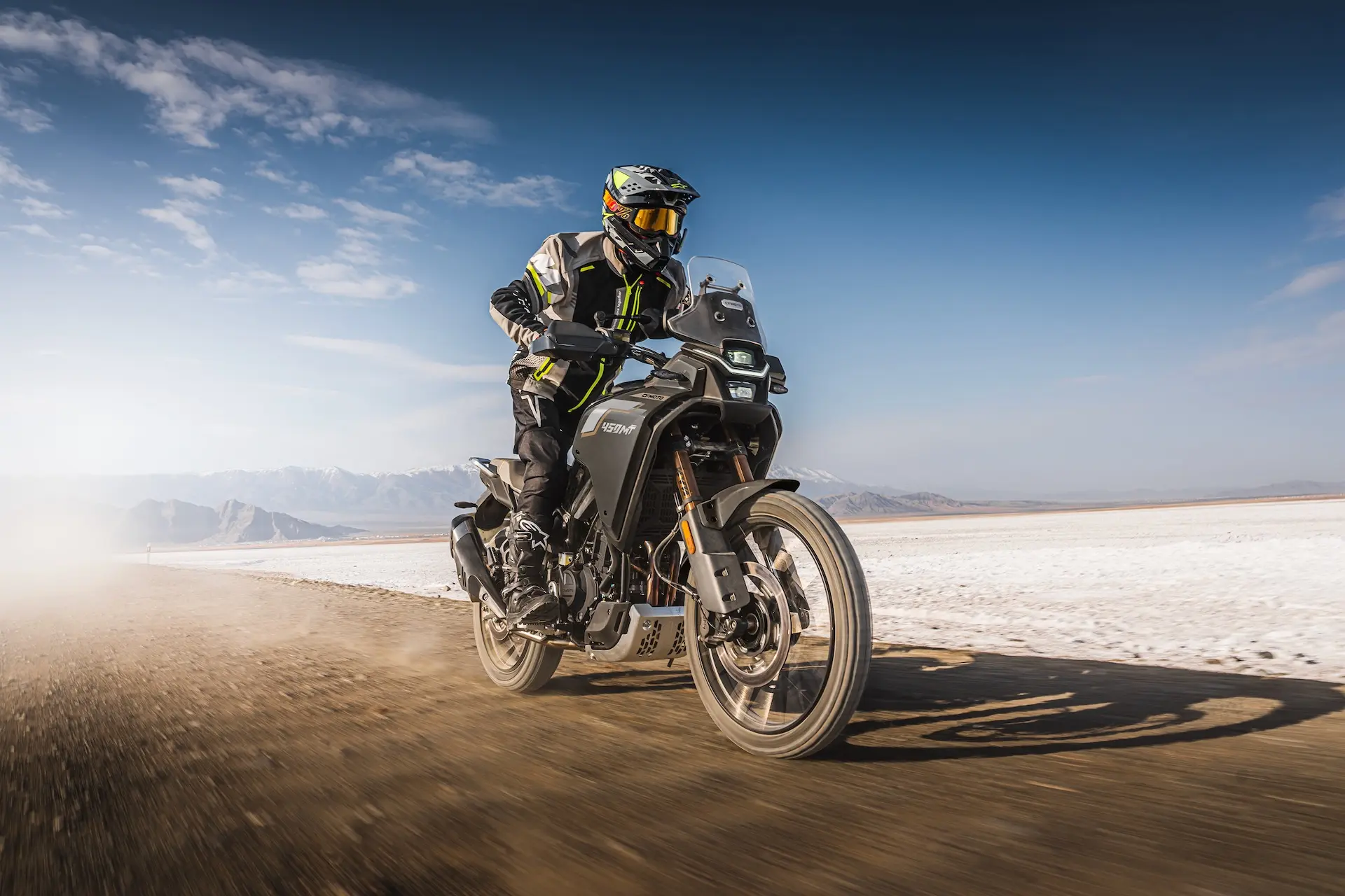A motorcyclist wearing safety gear rides a black 450MT adventure motorcycle on a dirt road, with mountains and a blue sky in the background.
