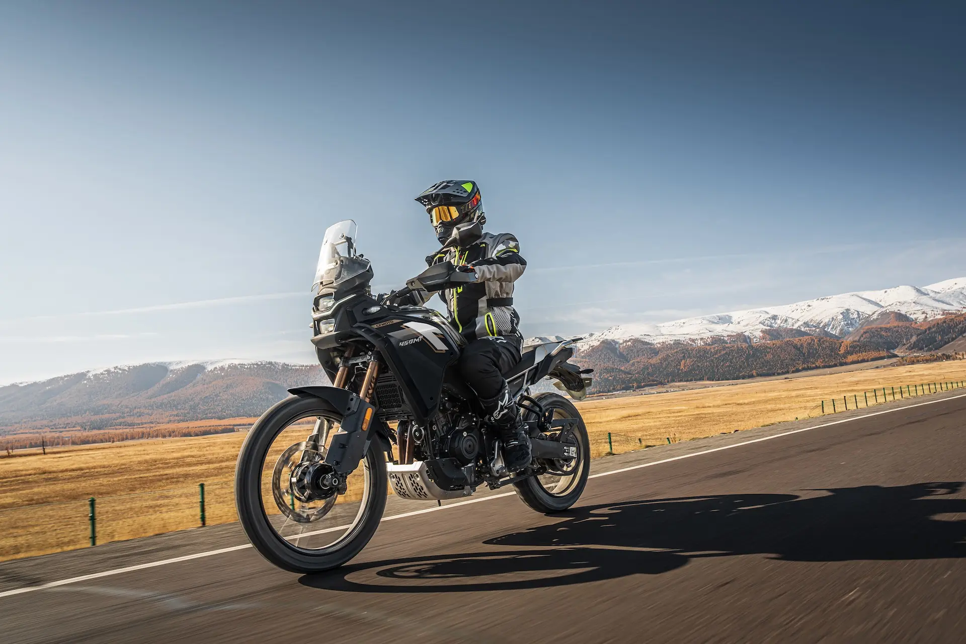 A motorcyclist wearing protective gear rides a 450MT adventure motorcycle on a paved road through a rural landscape with mountains in the background.