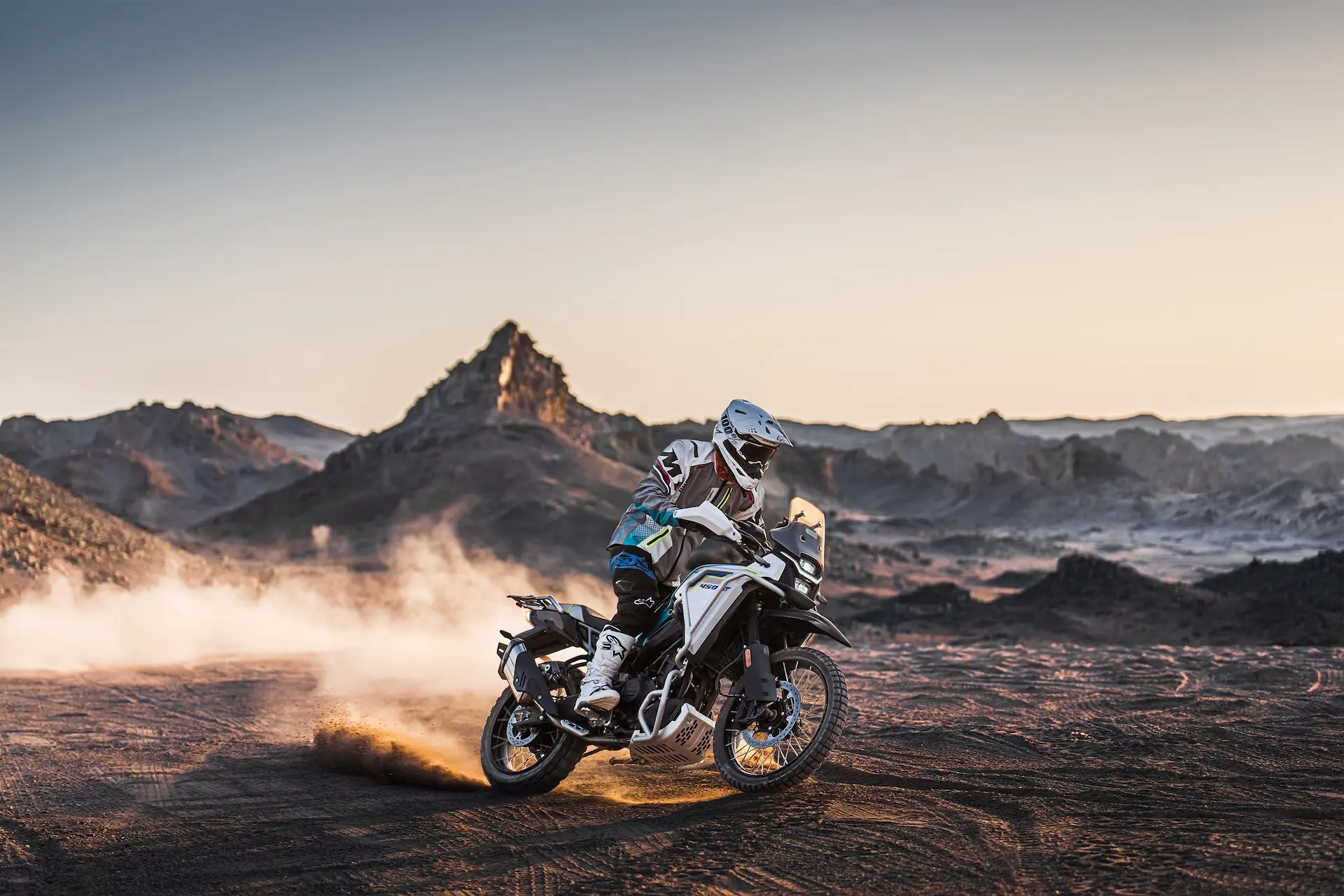A motorcyclist in full gear rides a 450MT adventure motorcycle on a dusty, rugged terrain with mountains in the background and a trail of dust behind the bike.