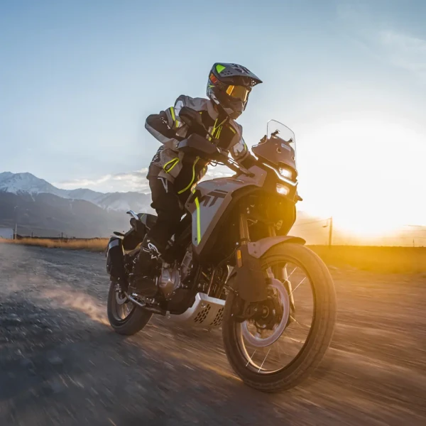 A person in full protective gear rides a motorcycle at speed on a gravel road with mountains and the sun in the background, showcasing an ideal adventure for new riders seeking excitement and skill-building.