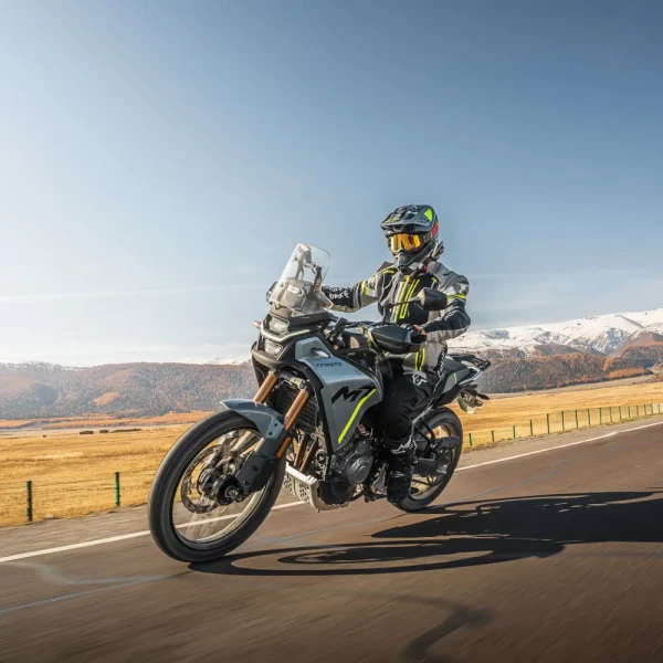 A person wearing full motorcycle gear enjoys leisure riding on an adventure motorcycle along a paved road, surrounded by scenic mountains and open fields.