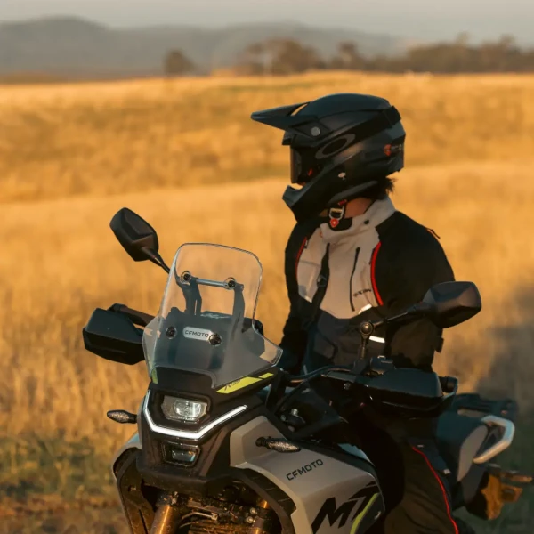 A person wearing motorcycle gear and helmet sits on a CF MOTO MT, one of the ideal first motorcycles for new riders, in a dry grassy field with hills in the background.