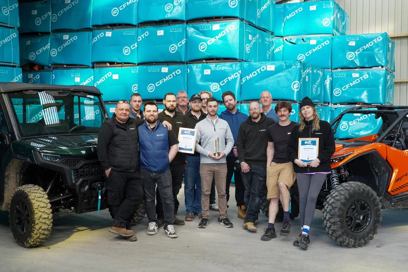 A group of people pose indoors between two off-road vehicles, holding certificates and a trophy, with stacked blue CFMOTO boxes in the background—celebrating their achievements on challenging motorcycle routes.