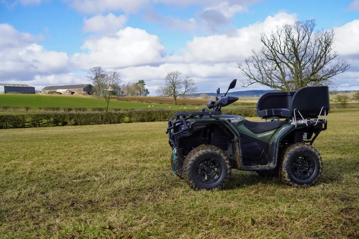 A green CFMOTO CForce 520 ATV is parked on a grassy field, framed by rural landscape, trees, and farm buildings under a partly cloudy sky.