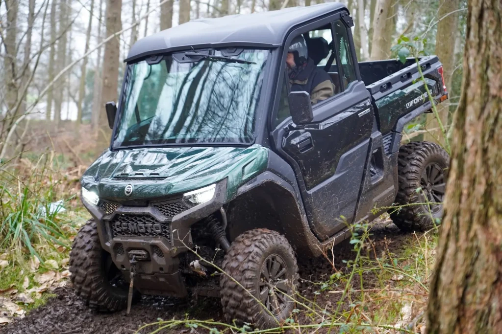 A green farm quad navigates through a muddy forest trail, surrounded by trees and underbrush.