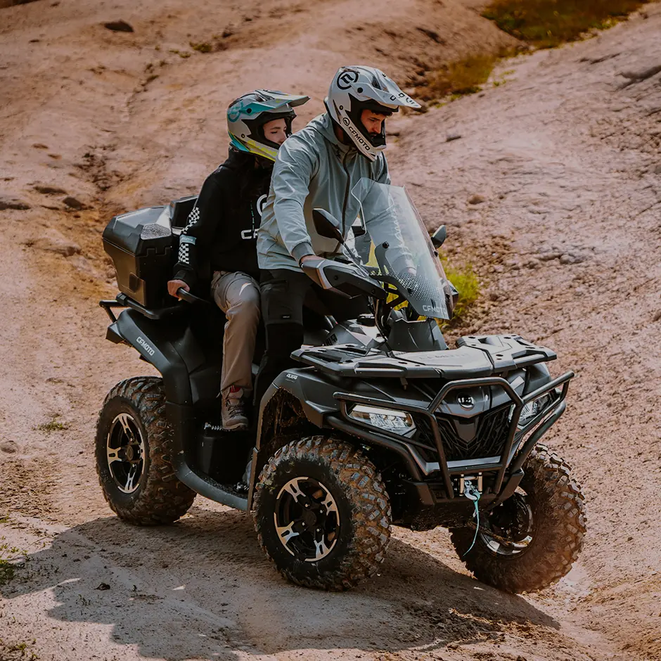 Two people wearing helmets ride a CFORCE 625 TOURING OVERLAND all-terrain vehicle (ATV) on a dirt path outdoors.