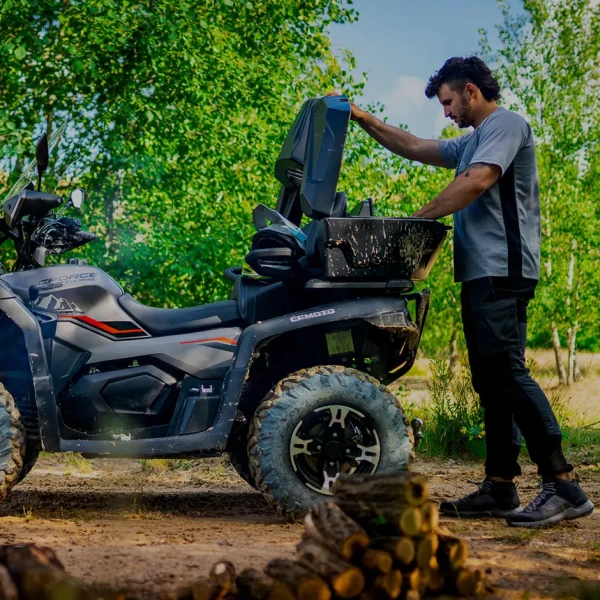 A man stands next to a CFORCE 625 TOURING OVERLAND ATV, lifting the rear storage compartment open, with a stack of firewood in the foreground and green trees in the background.