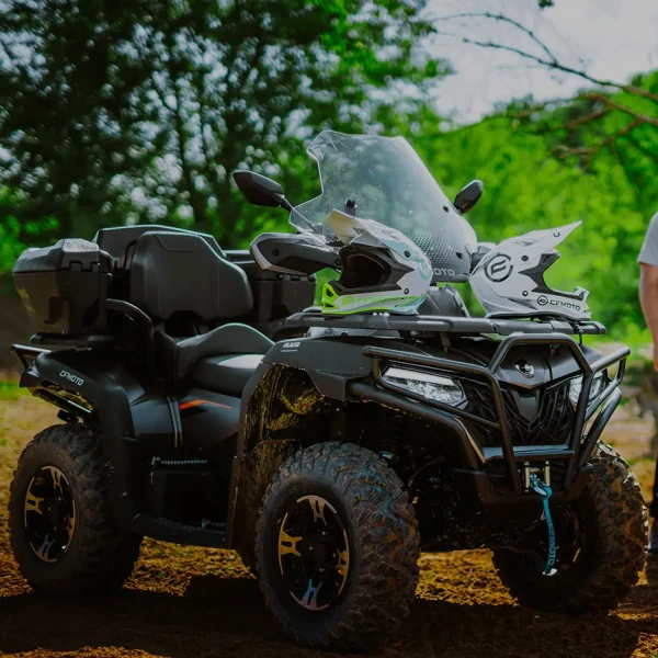 A black CFORCE 625 TOURING OVERLAND all-terrain vehicle with two helmets on the handlebars is parked outdoors on dirt, with green trees in the background.