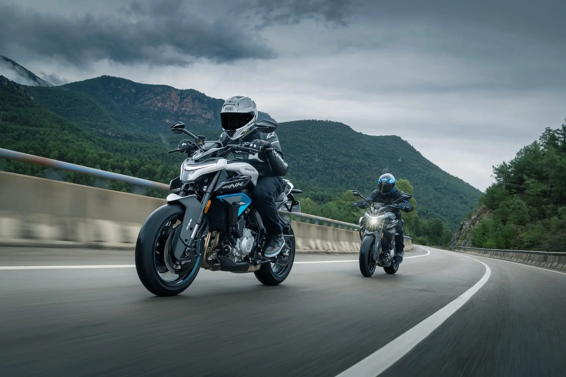 Two motorcyclists ride at speed on a curved mountain road, both astride their 125NK bikes, with trees and hills in the background under a cloudy sky.