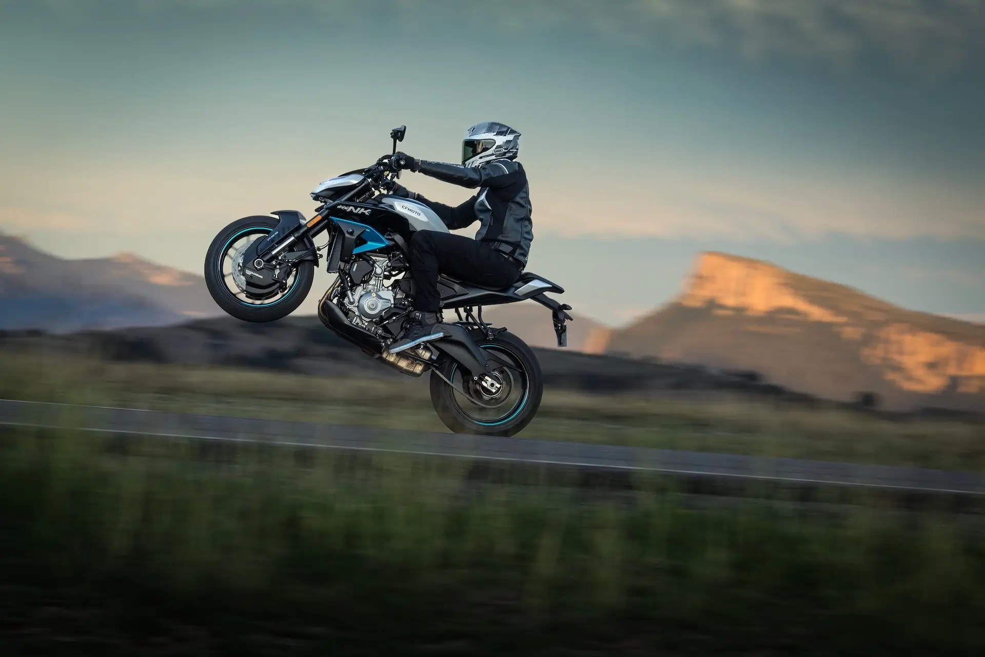 A person wearing protective gear performs a wheelie on a 125NK motorcycle on a road with mountains in the background.