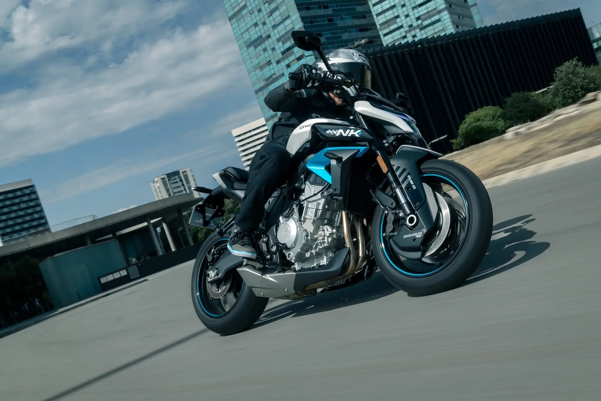 A person wearing black gear and a helmet rides a modern 125NK sport motorcycle on a city street, with tall buildings in the background.