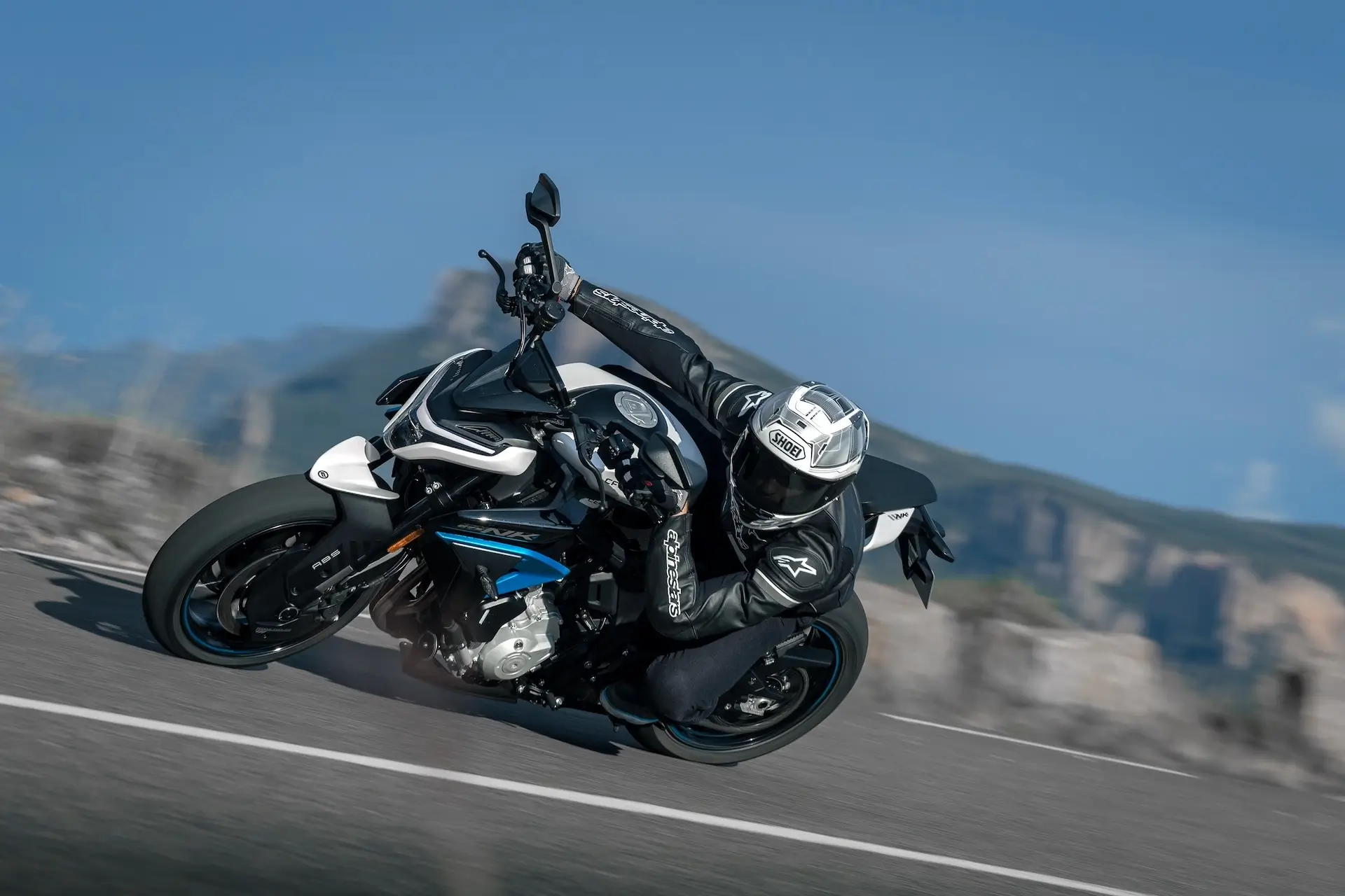 A motorcyclist wearing full protective gear leans into a turn on a blue and white 125NK sport motorcycle on a paved road with mountains in the background.