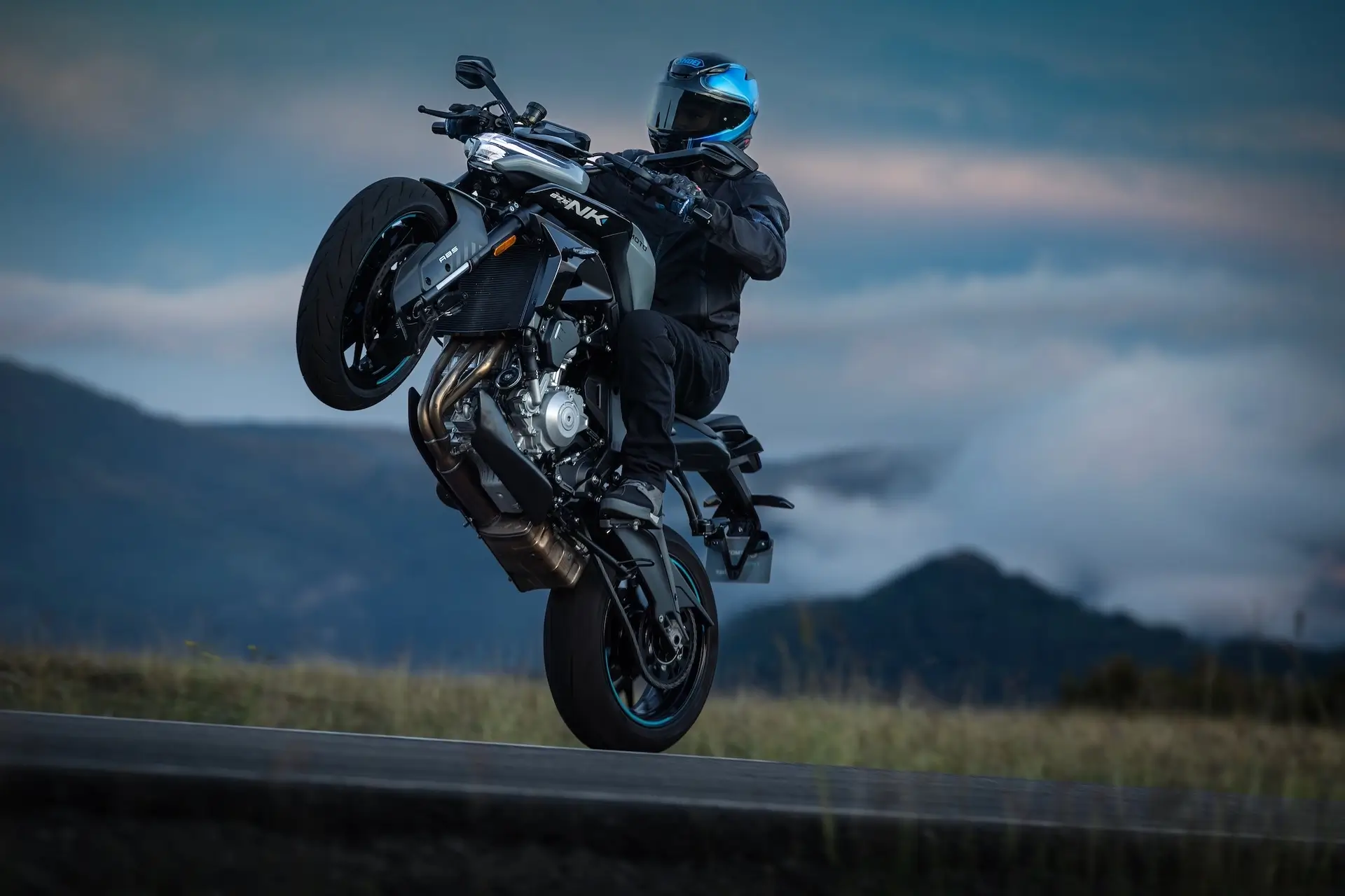 A motorcyclist in full gear and a blue helmet performs a wheelie on a 125NK sport bike on an outdoor road with mountains in the background.