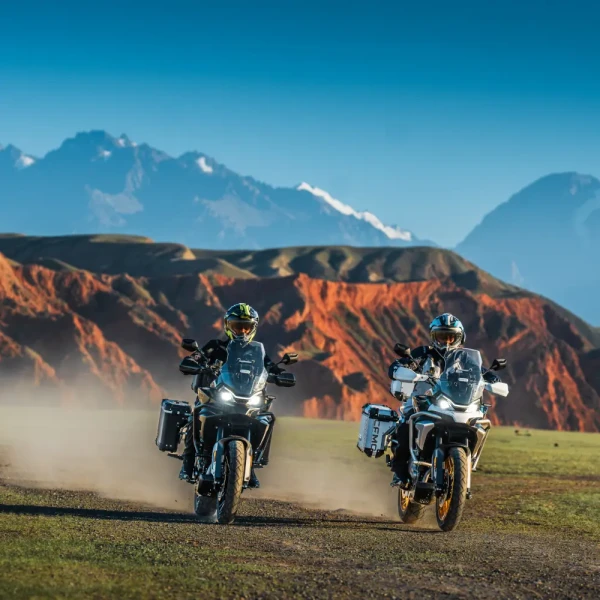 Two motorcyclists ride 700MT adventure bikes on a dirt path through a scenic landscape with mountains and red rock formations in the background.