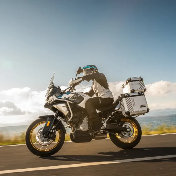 A person rides a white 700MT adventure motorcycle with storage boxes on a paved road near a body of water under a partly cloudy sky.