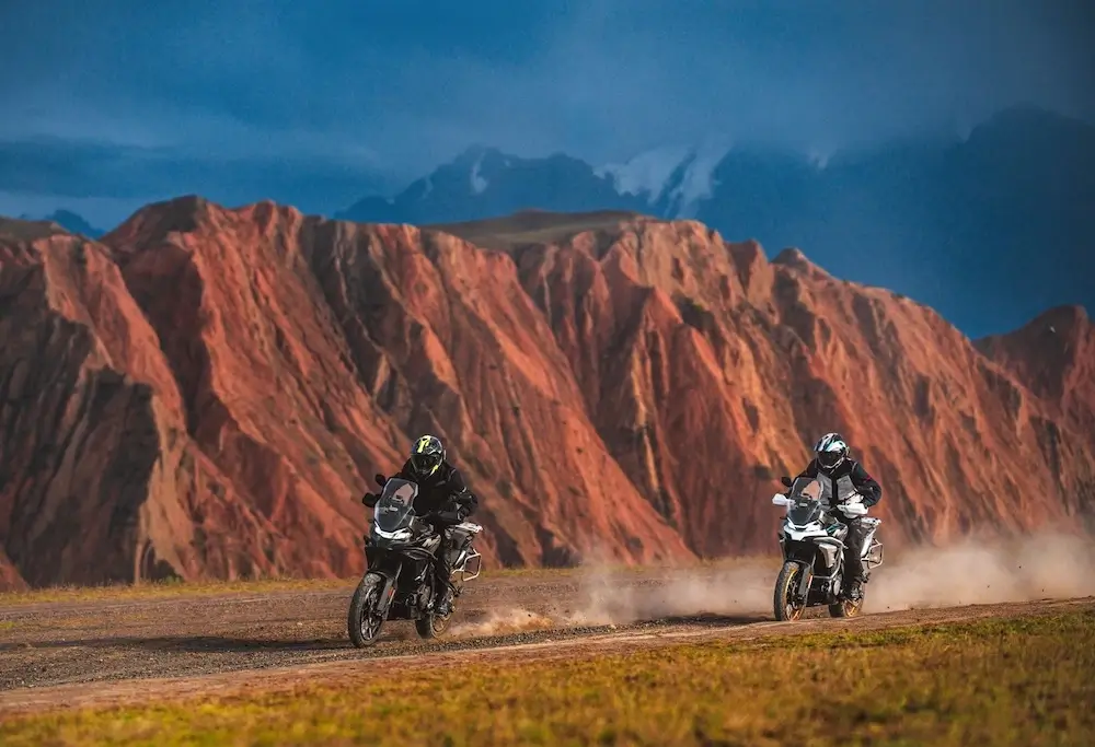 Two motorcyclists ride on a dirt road, kicking up dust as their CFMOTO T-Box tech tracks their adventure, with dramatic red rock formations and a cloudy sky in the background.
