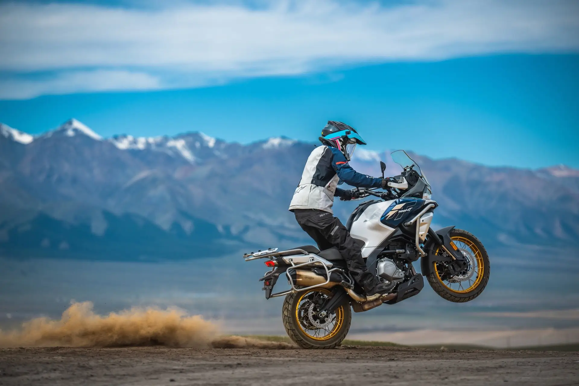 A motorcyclist in protective gear performs a wheelie on a 450MT along a dirt road, with mountains and blue sky in the background.