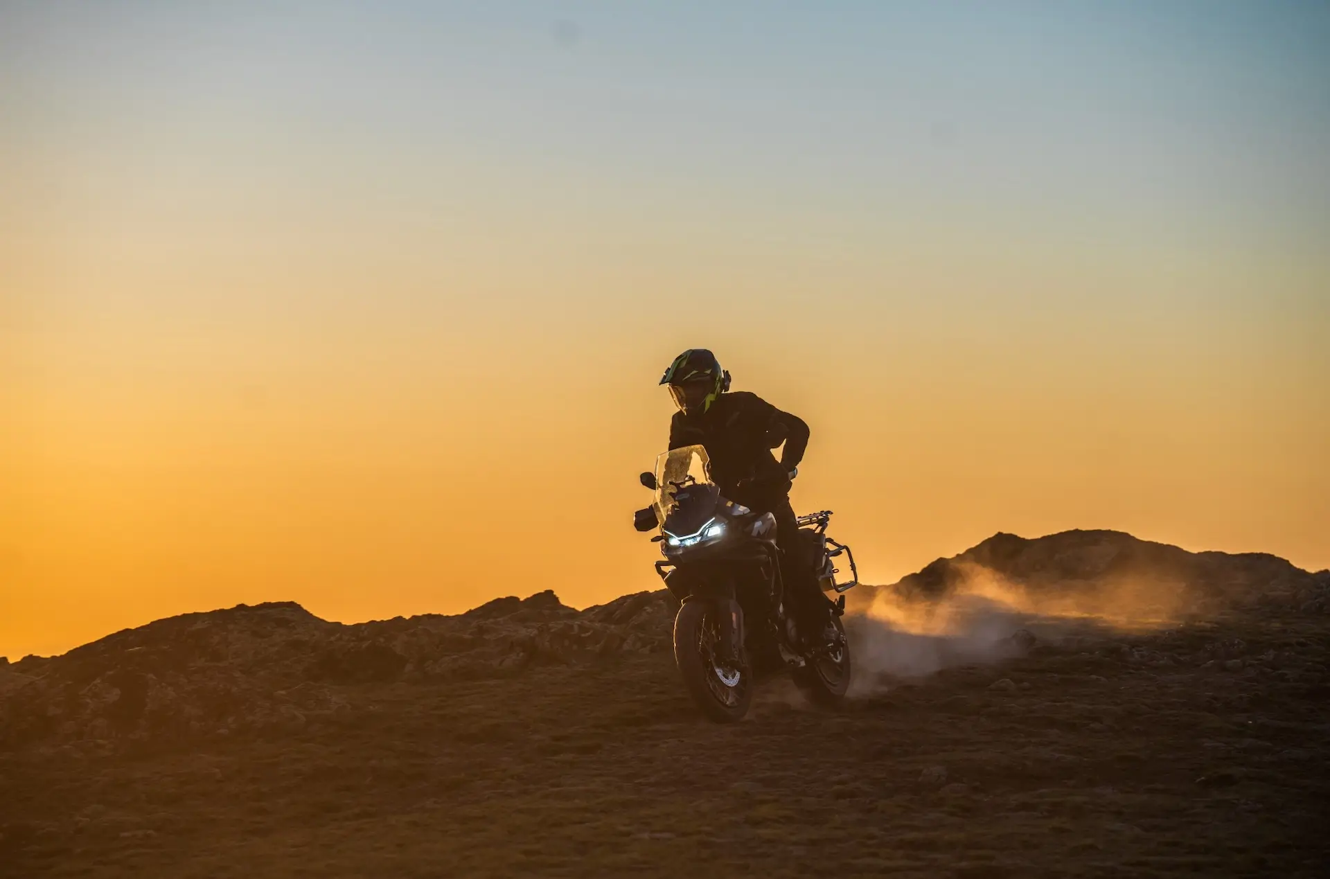 A person rides a 450MT motorcycle on rough terrain at sunset, kicking up dust with rocky hills silhouetted in the background.