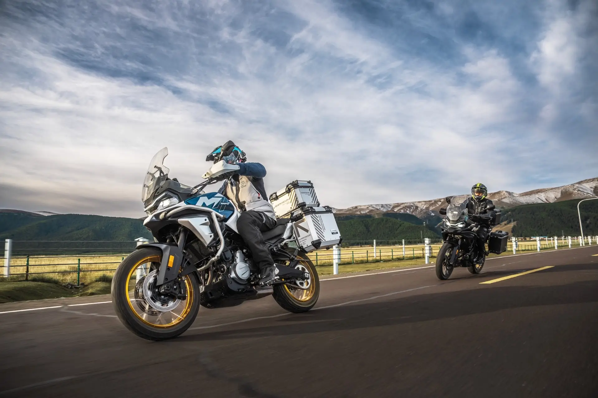 Two motorcyclists ride 450MT adventure touring bikes on an open road, framed by rugged mountains and a partly cloudy sky in the background.