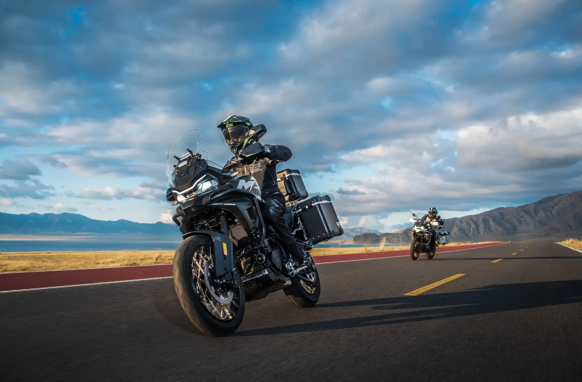 Two motorcyclists ride 450MT adventure touring bikes on an open road with mountains and partly cloudy skies in the background.