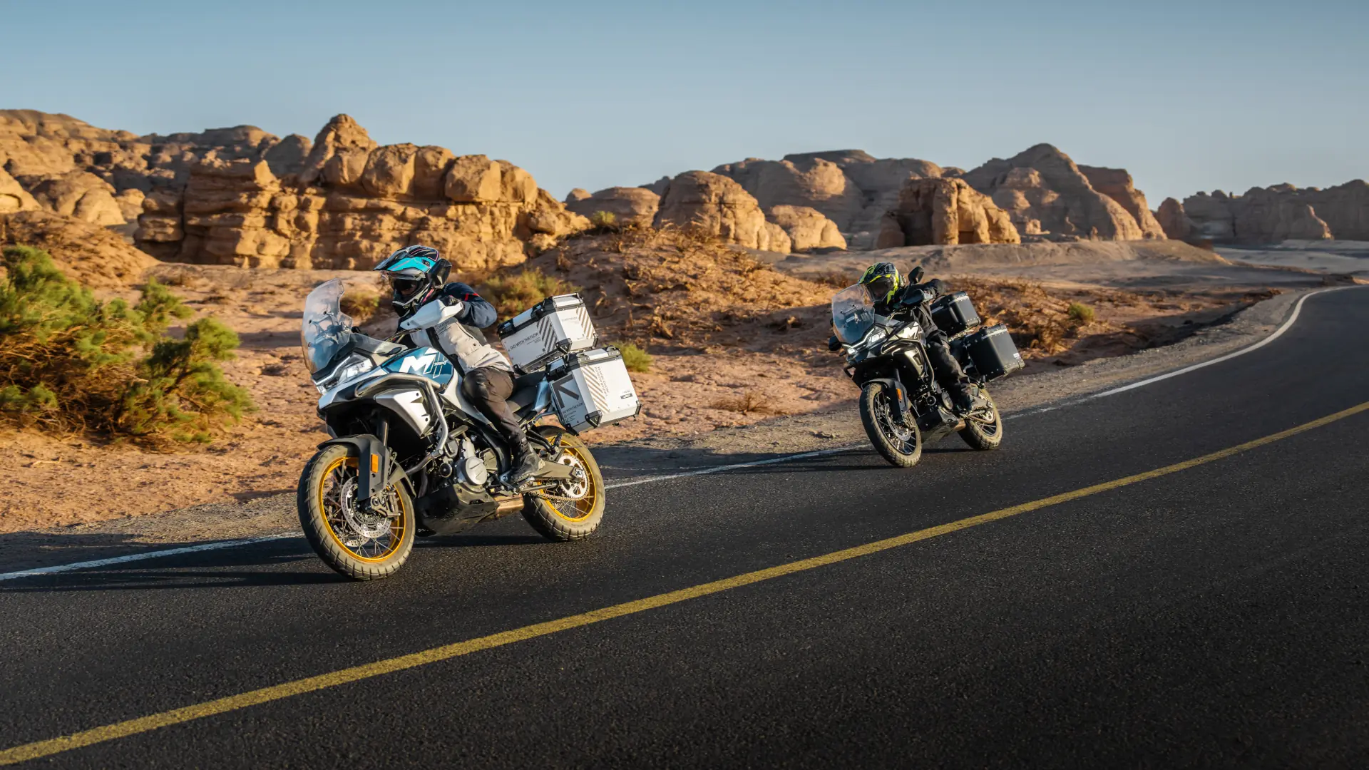 Two motorcyclists ride 450MT adventure bikes with luggage cases along a paved desert road, rocky formations visible in the background under a clear sky.