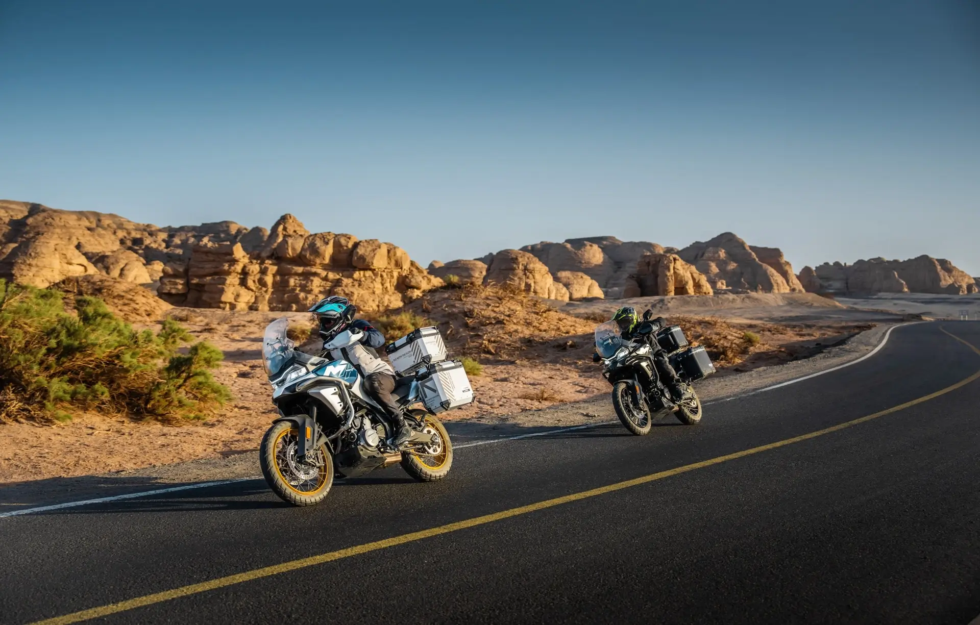 Two motorcyclists ride 450MT adventure bikes on a winding desert road, framed by rocky formations and a clear blue sky in the background.
