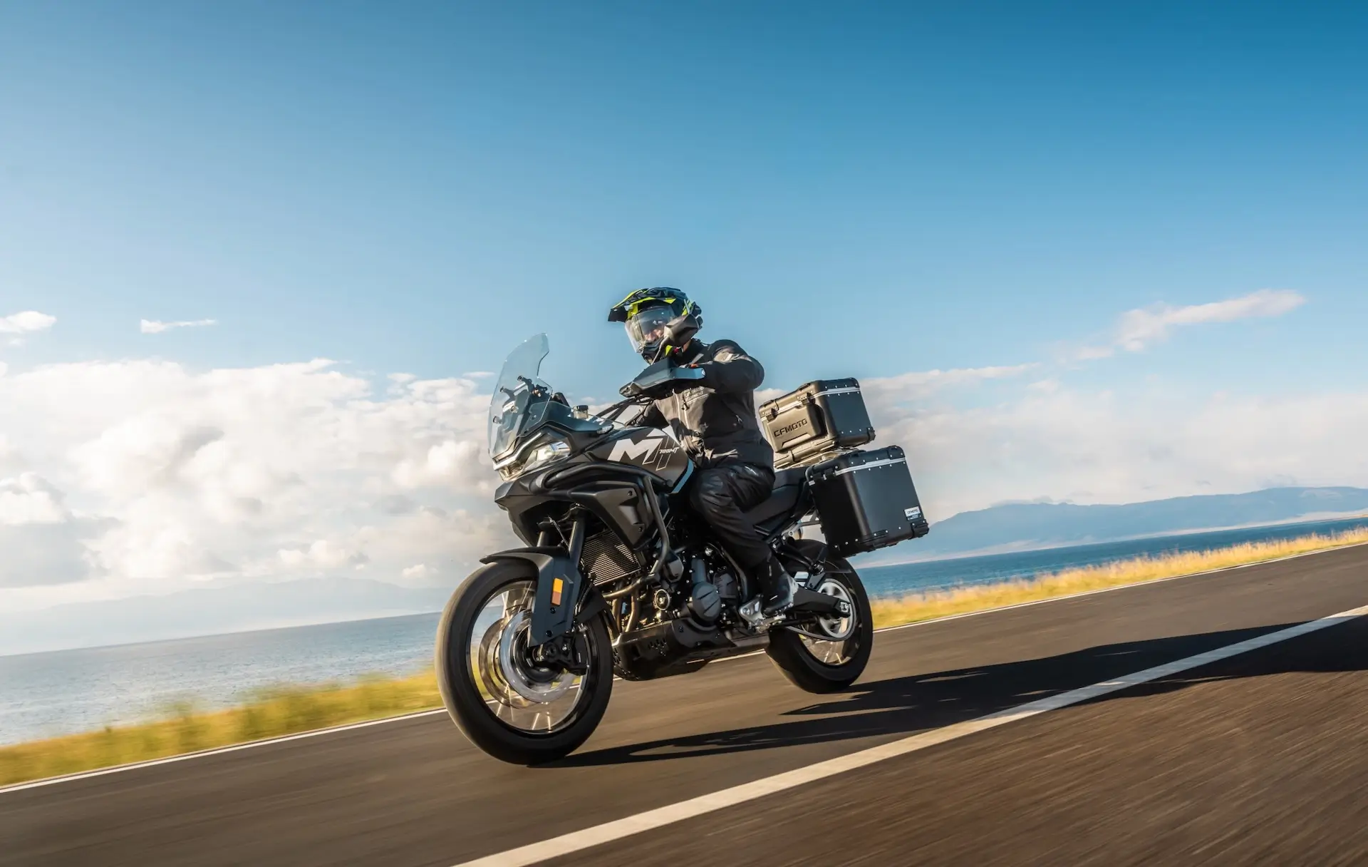A motorcyclist rides a 450MT touring bike with side panniers and top case on an open road beside a body of water under a clear blue sky.