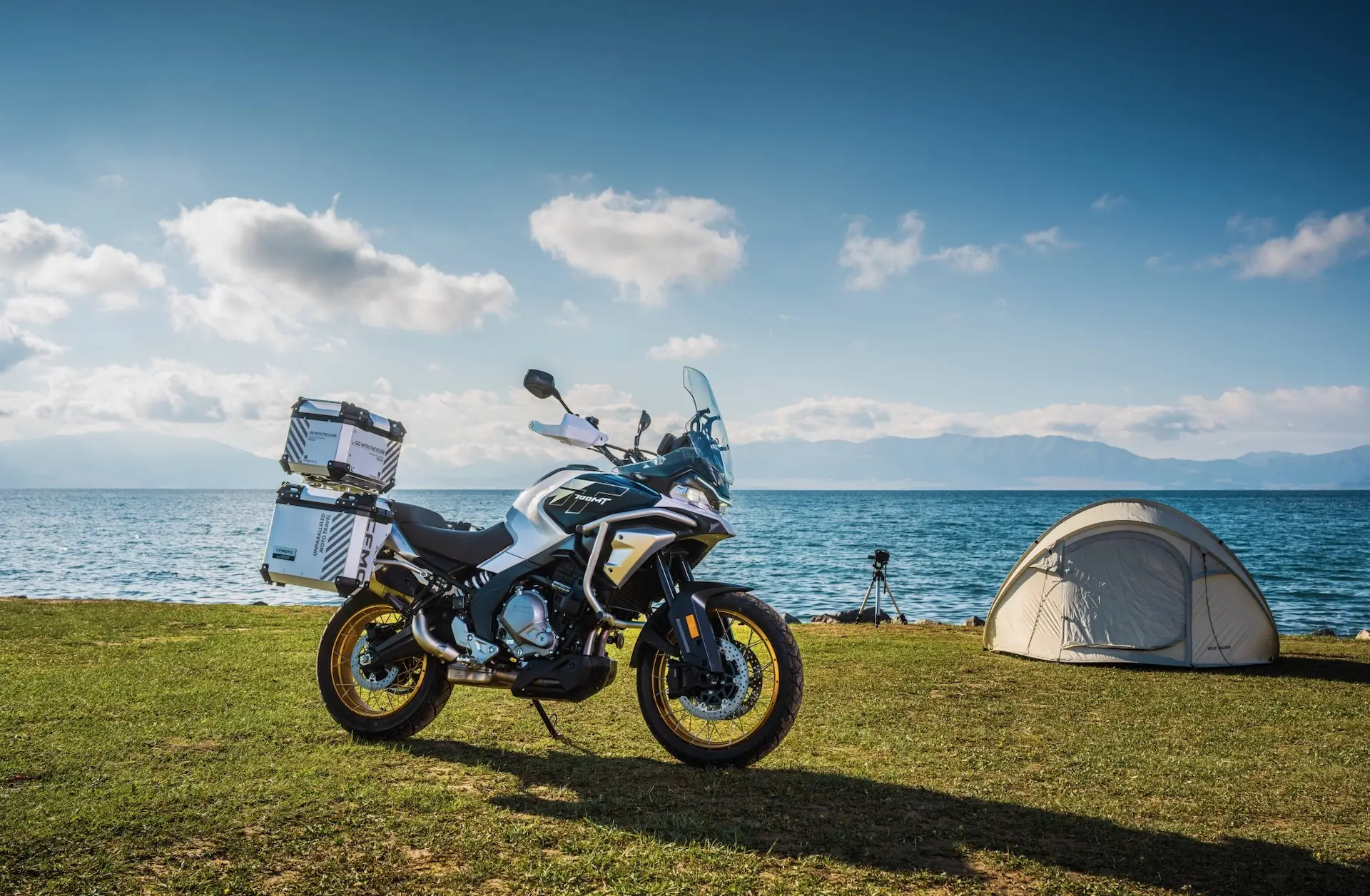 A 450MT touring motorcycle is parked on grass near a tent by a lake, with mountains in the distance under a blue sky with scattered clouds.