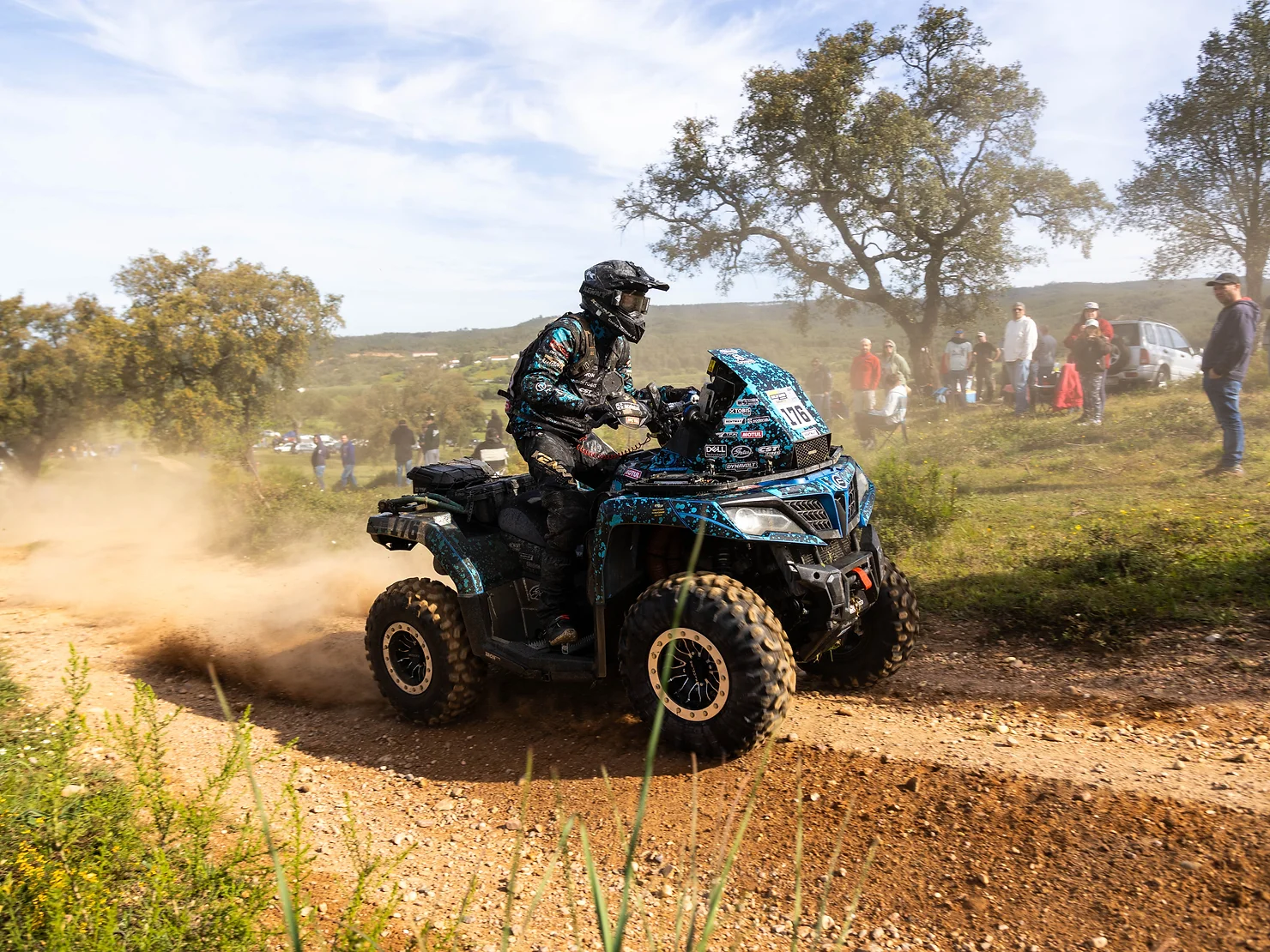 A person races an all-terrain vehicle (ATV) on a dirt path in Greece, creating clouds of dust. Spectators watch intently in the background as if witnessing a thrilling CFMOTO Thunder Racing Team event.