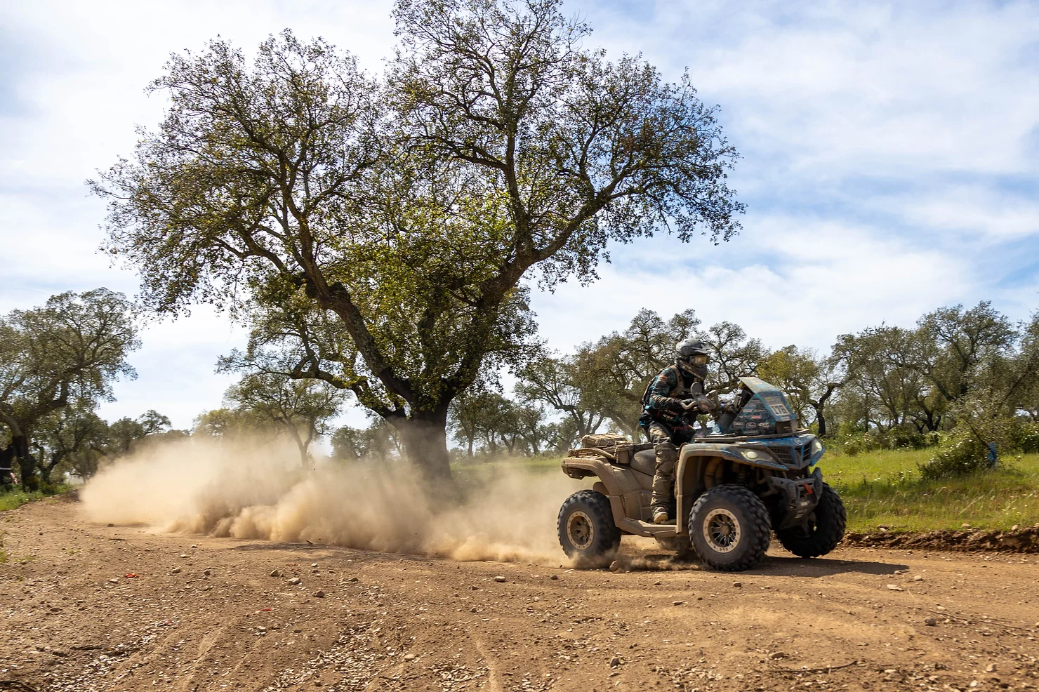 A person riding an ATV on a dirt path, kicking up a trail of dust, with trees and a blue sky reminiscent of Greece's scenic beauty. It's like watching the CFMOTO Thunder Racing Team in action, embodying the same spirit of adventure and precision.