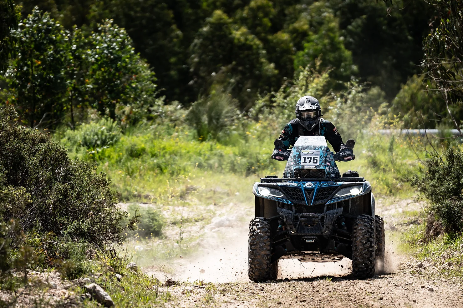 A person rides an ATV on a dirt trail, surrounded by greenery and trees, with dust kicked up behind them, embodying the spirit of the CFMOTO Thunder Racing Team.