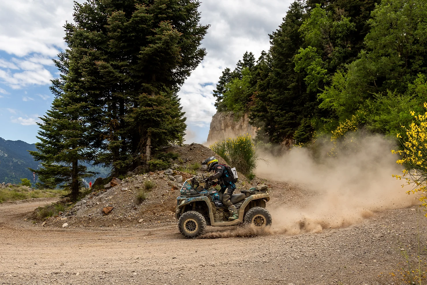 On the second day, a person drove an ATV on a dusty dirt trail in Greece, creating swirling clouds under a partly cloudy sky, surrounded by lush trees and shrubs.