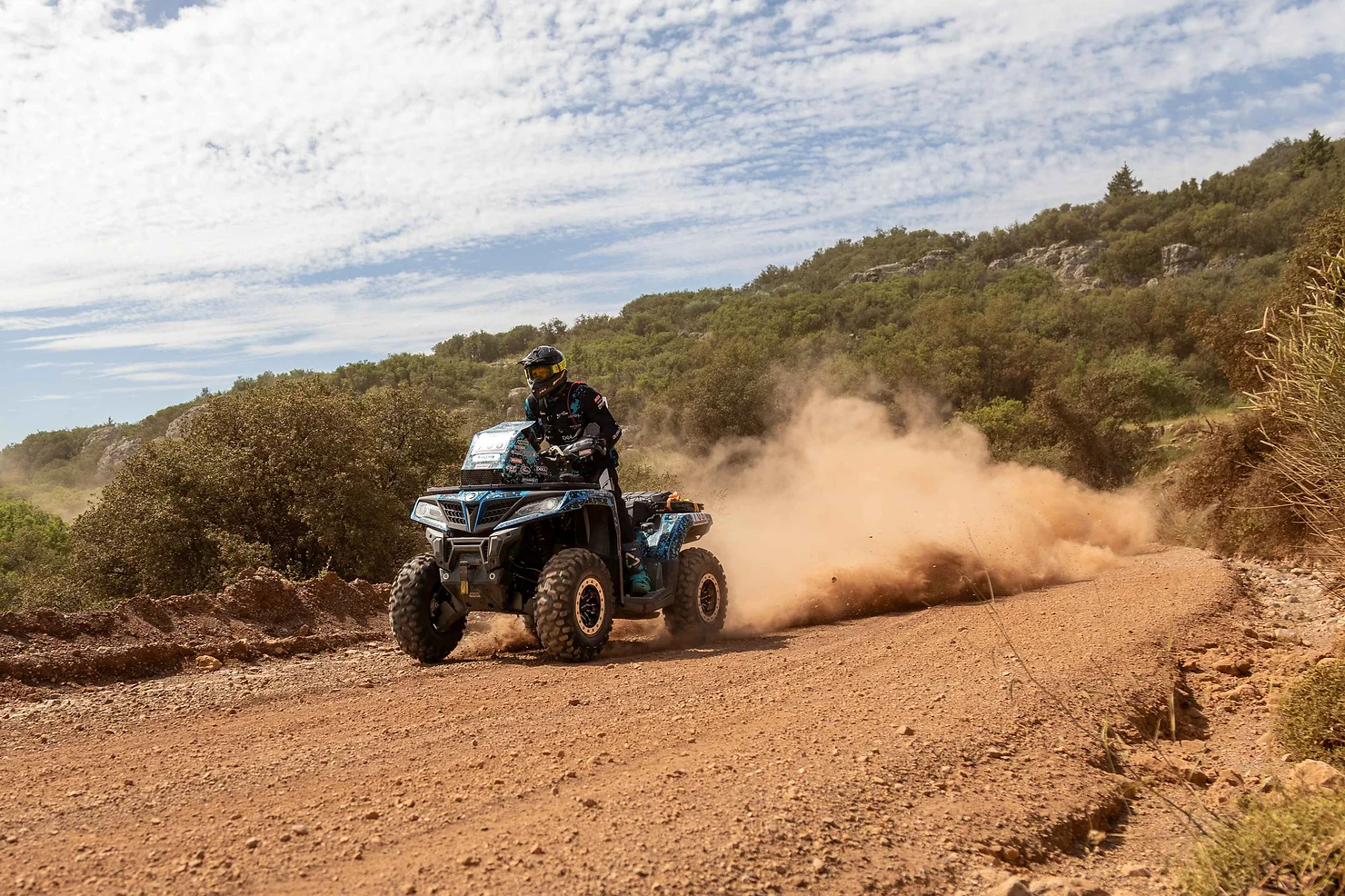 A person is rallying their ATV on a dusty, rural trail in Greece under a partly cloudy sky.