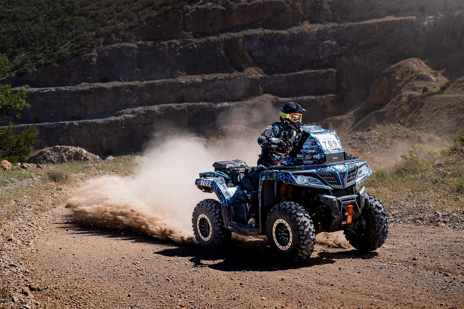 A person rides an ATV on a dirt trail, kicking up dust, with rocky hills in the background, reminiscent of a challenging day at the Greece rally.