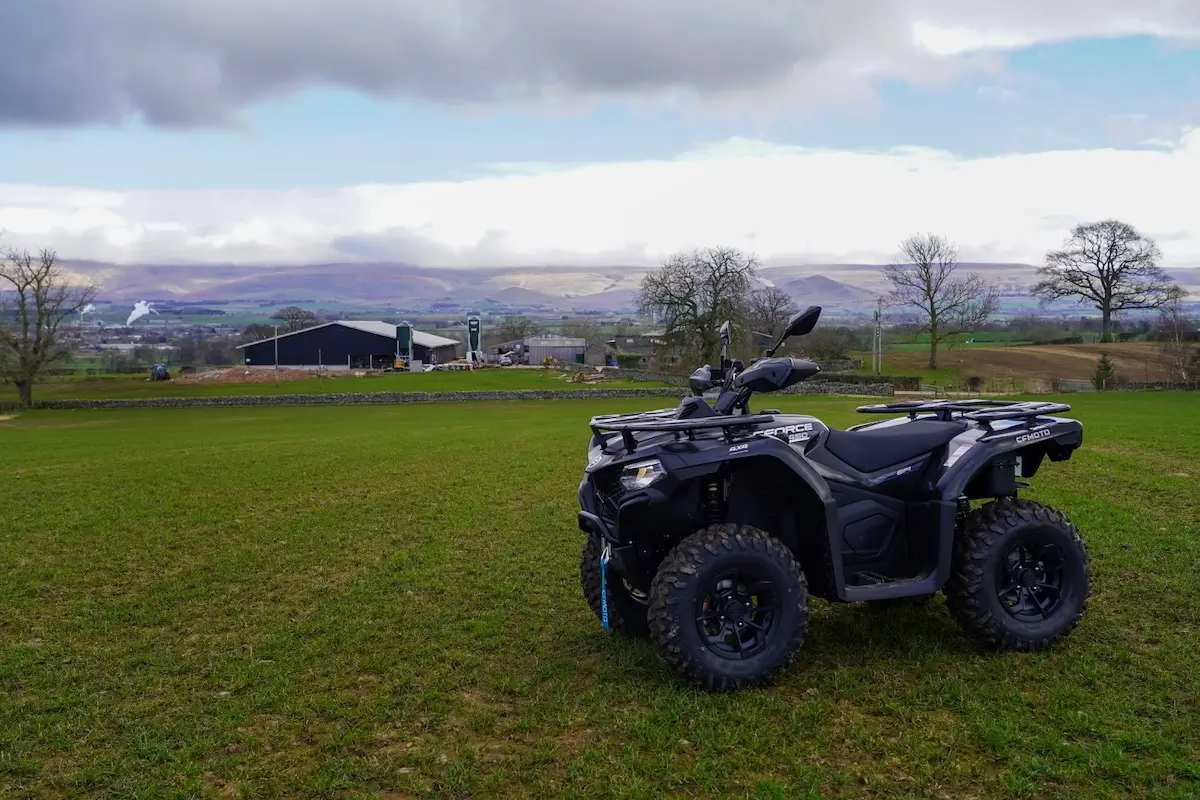 A black CForce 450 all-terrain vehicle is parked on a grassy field with a farm building and hills in the background under a cloudy sky.