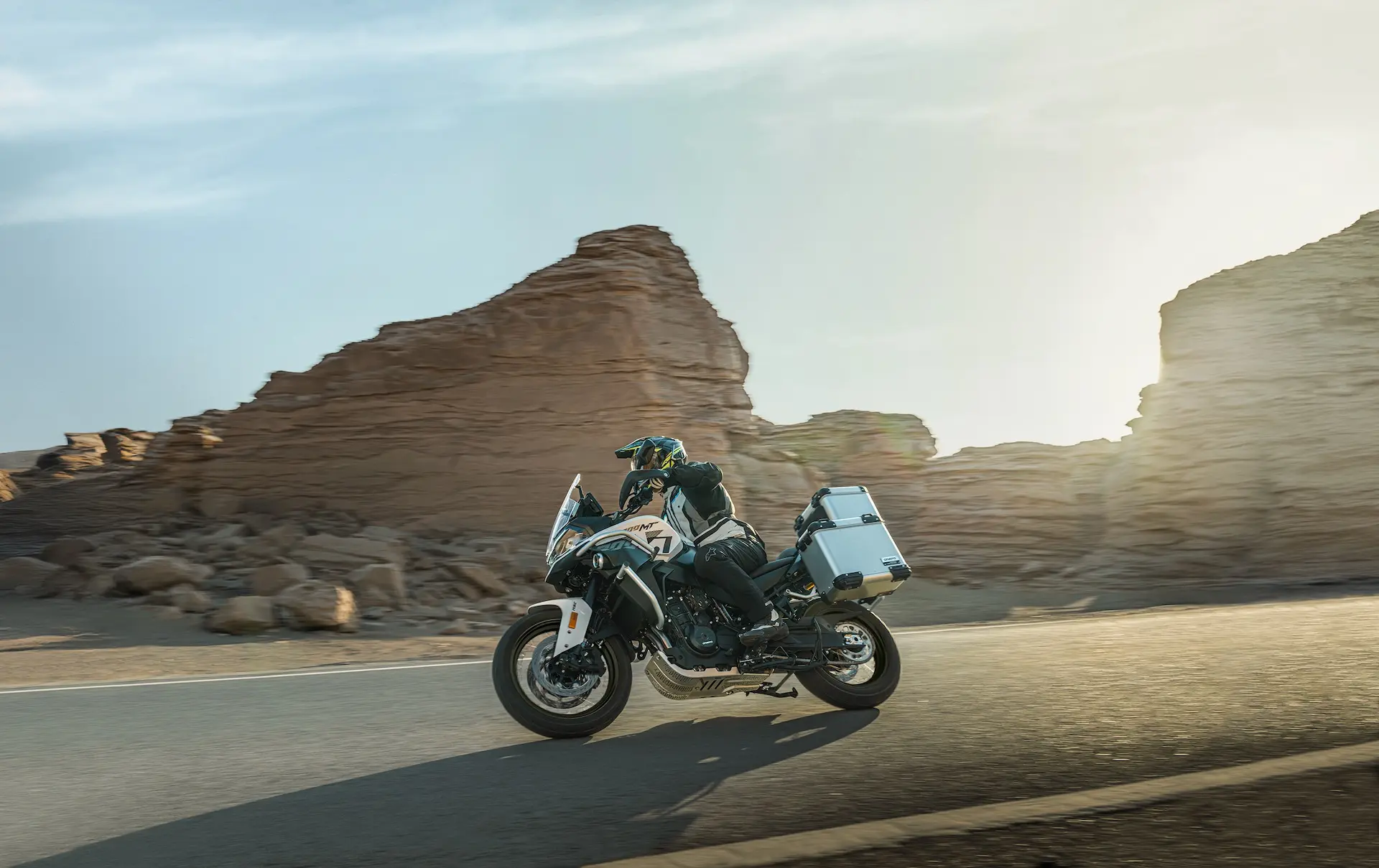 A person rides an 800MT-X touring motorbike on a tarmacked road through a rocky desert landscape with the sun low in the sky.