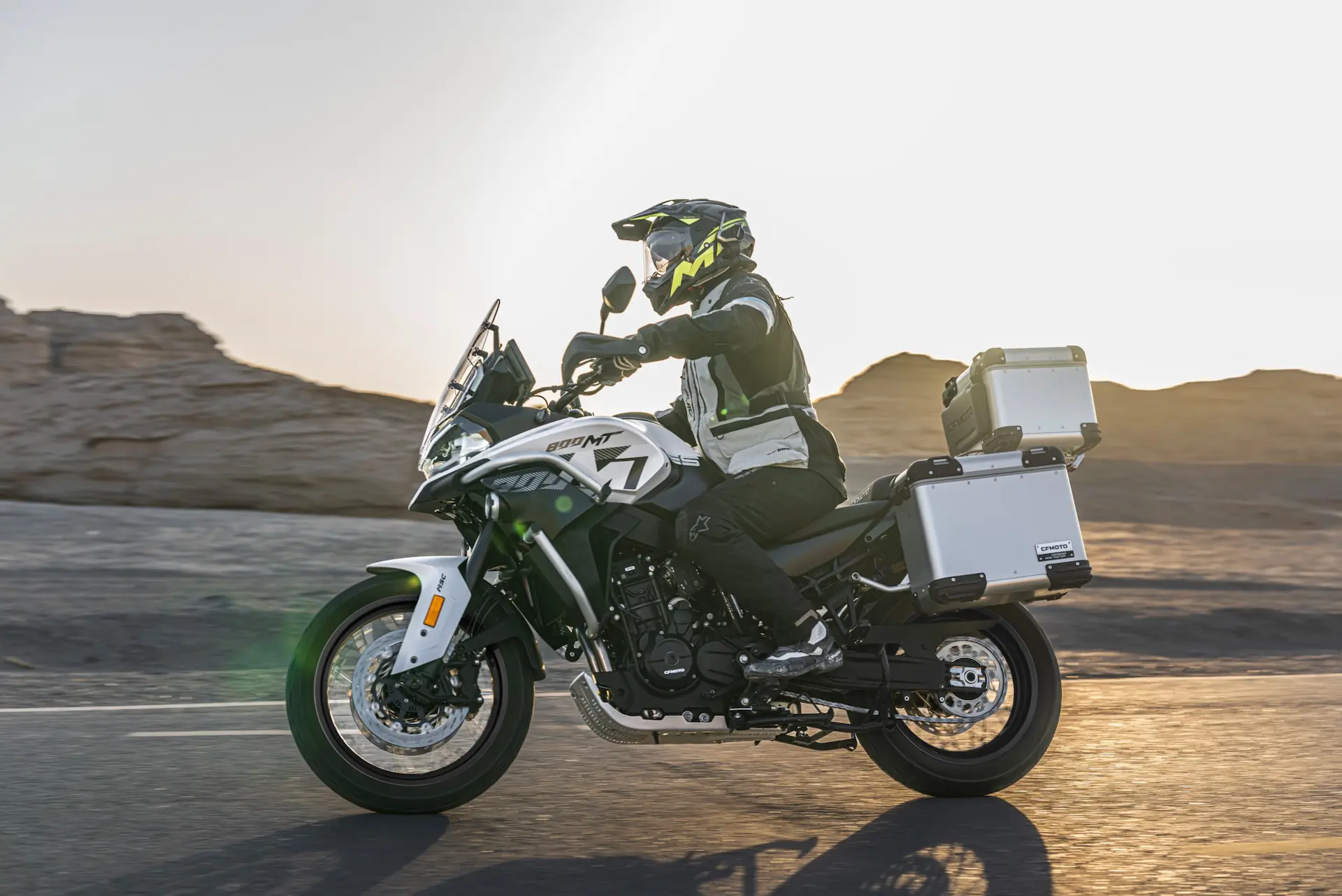A person in riding gear rides a white 800MT-X adventure motorbike with panniers on a tarmacked road through a desert landscape at sunset.