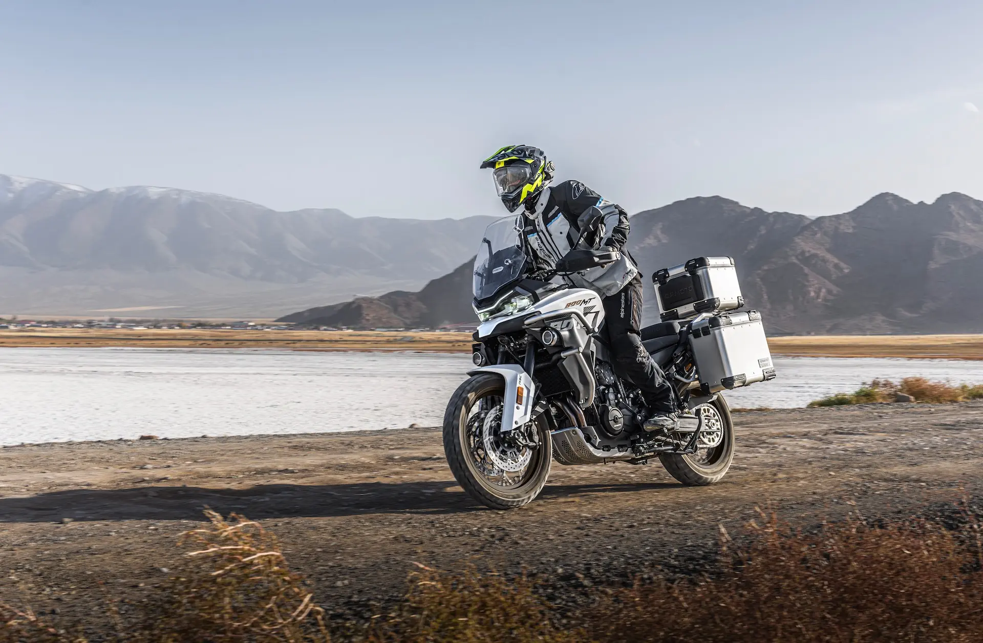 A motorcyclist wearing protective gear rides an 800MT-X adventure touring motorbike with side and rear storage boxes on a dirt road near mountains and a dry lakebed.
