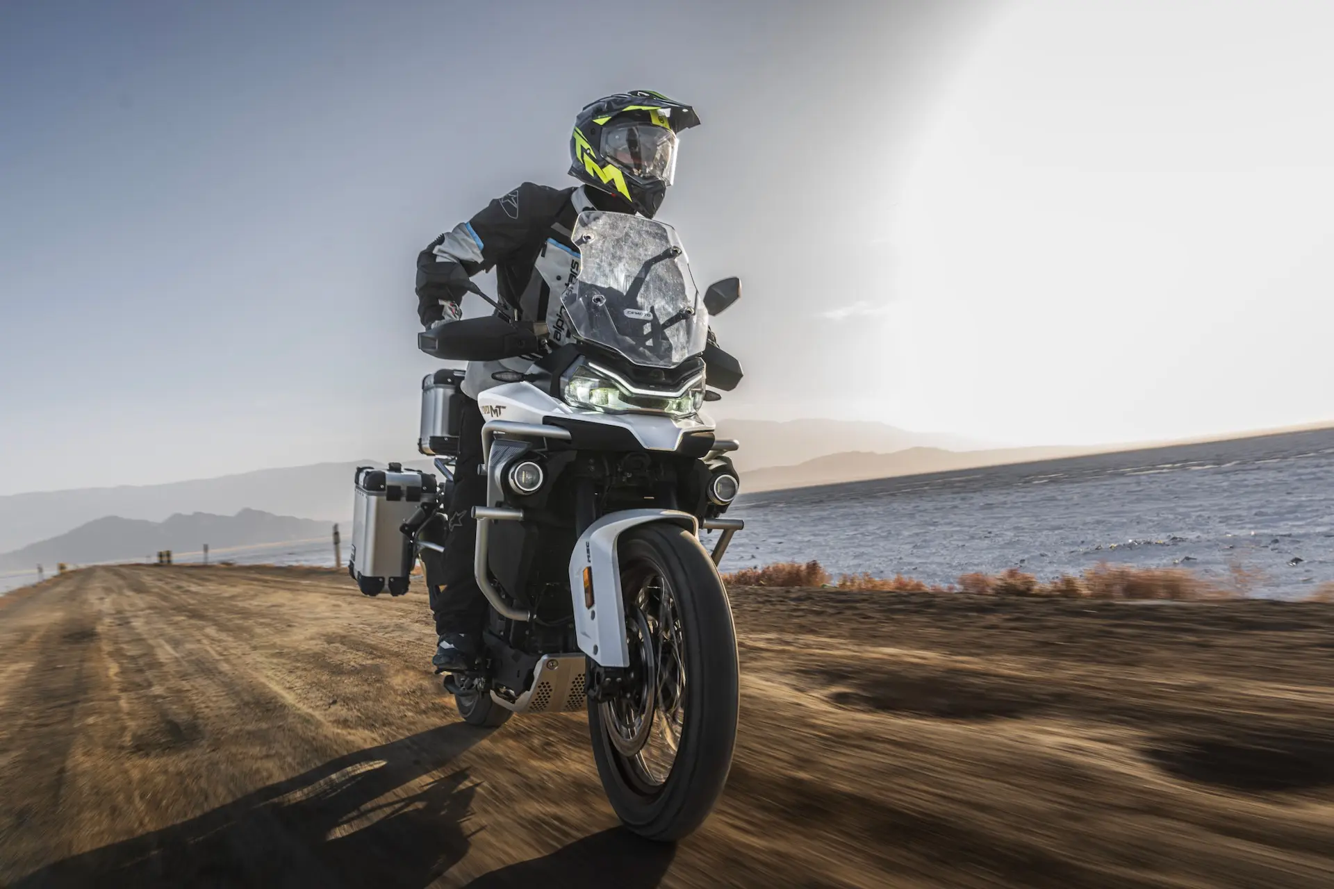 A motorcyclist wearing protective gear rides an 800MT-X adventure motorbike on a dirt road beside a body of water with mountains in the background.