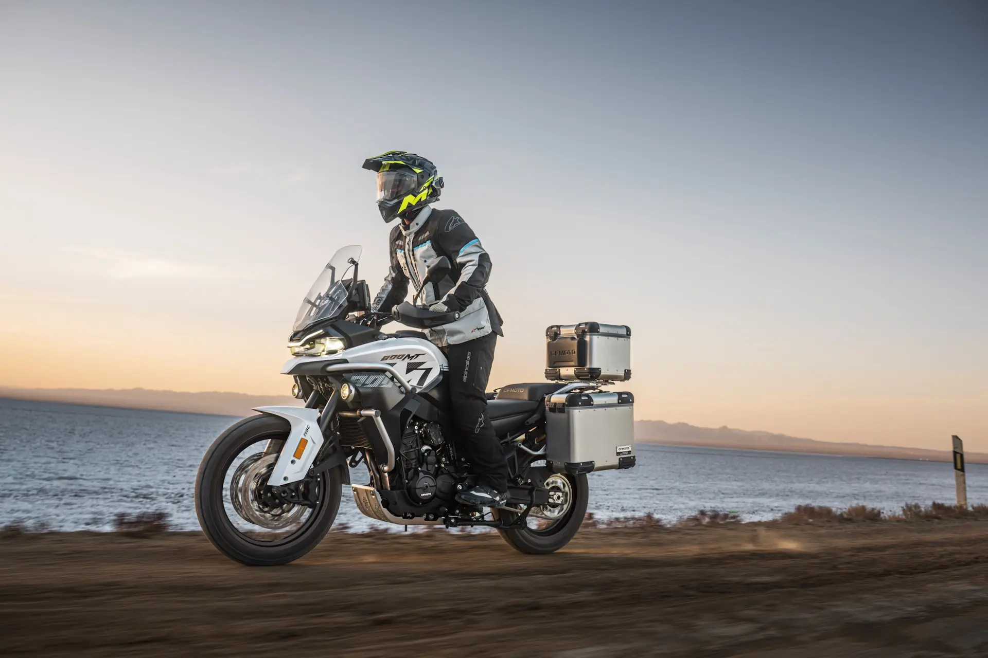 A person wearing motorcycle gear rides an 800MT-X adventure motorbike with panniers along a dirt track beside a body of water at sunset.