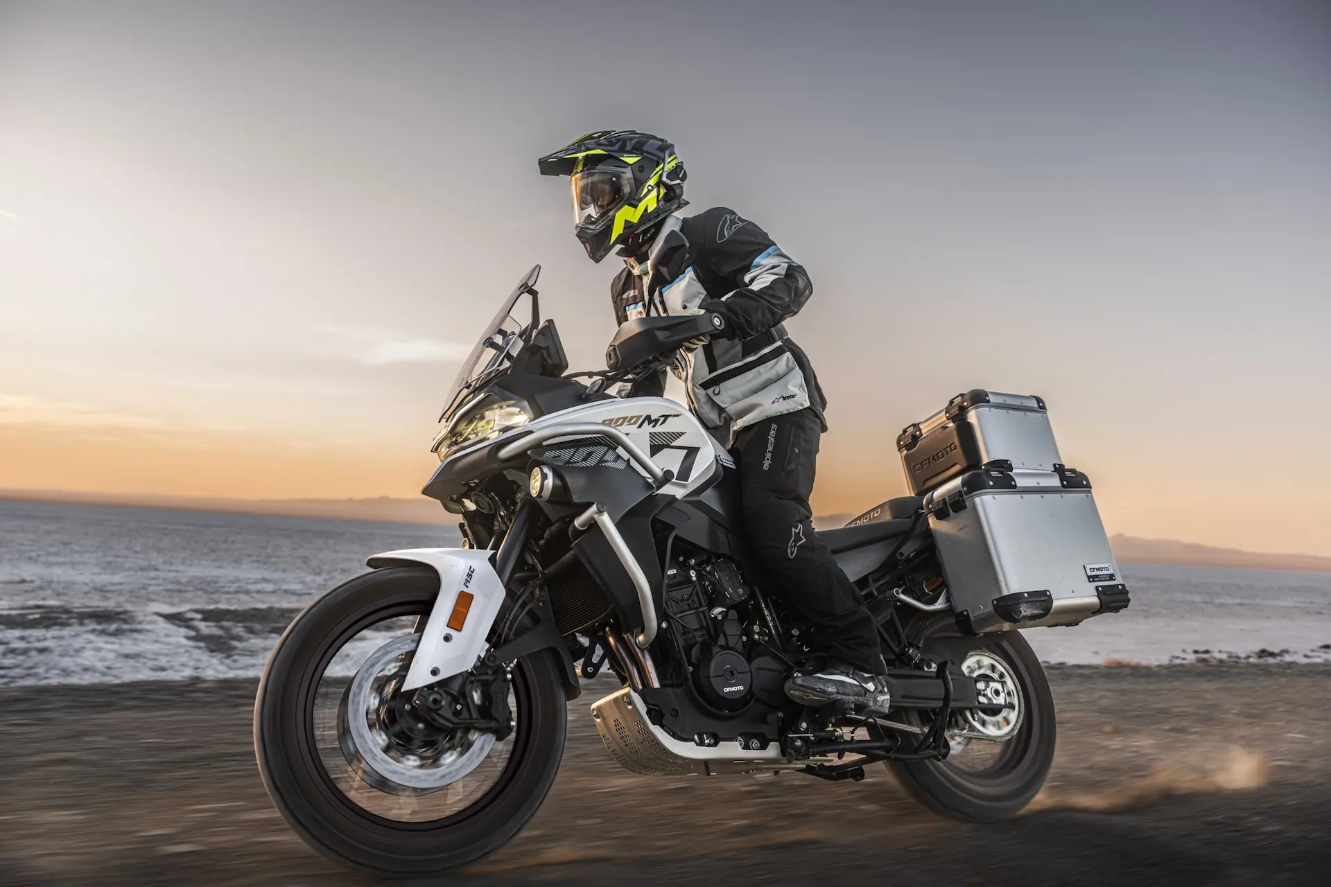 A person wearing protective gear rides an 800MT-X adventure motorbike with side and rear storage boxes on a dirt track near a body of water at sunset.