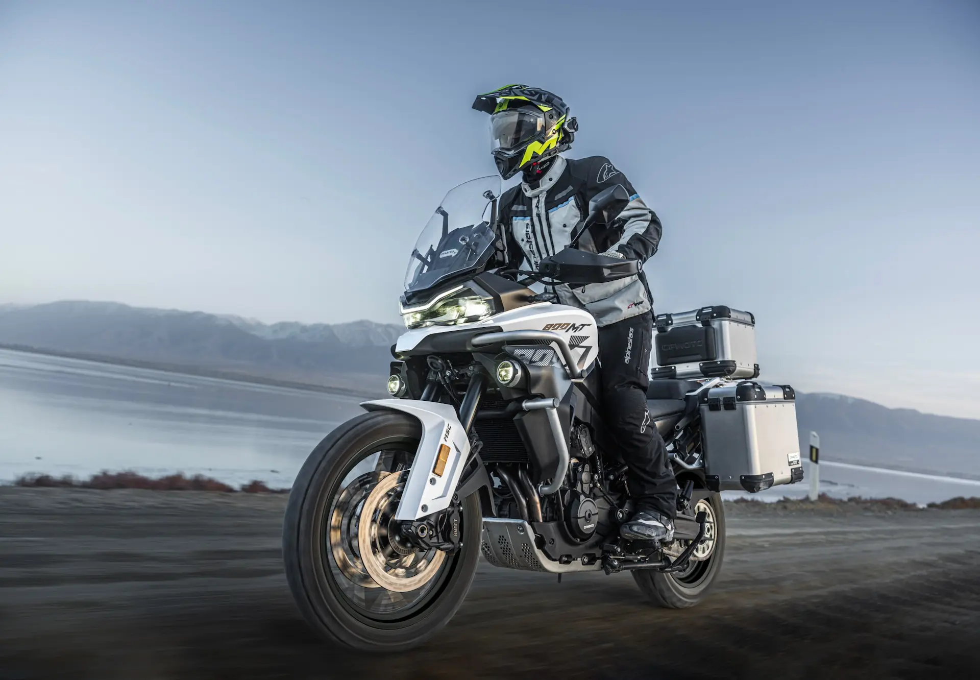 A person wearing riding kit and a helmet is riding an 800MT-X touring motorbike with panniers on a dirt track near a body of water, with mountains in the background.