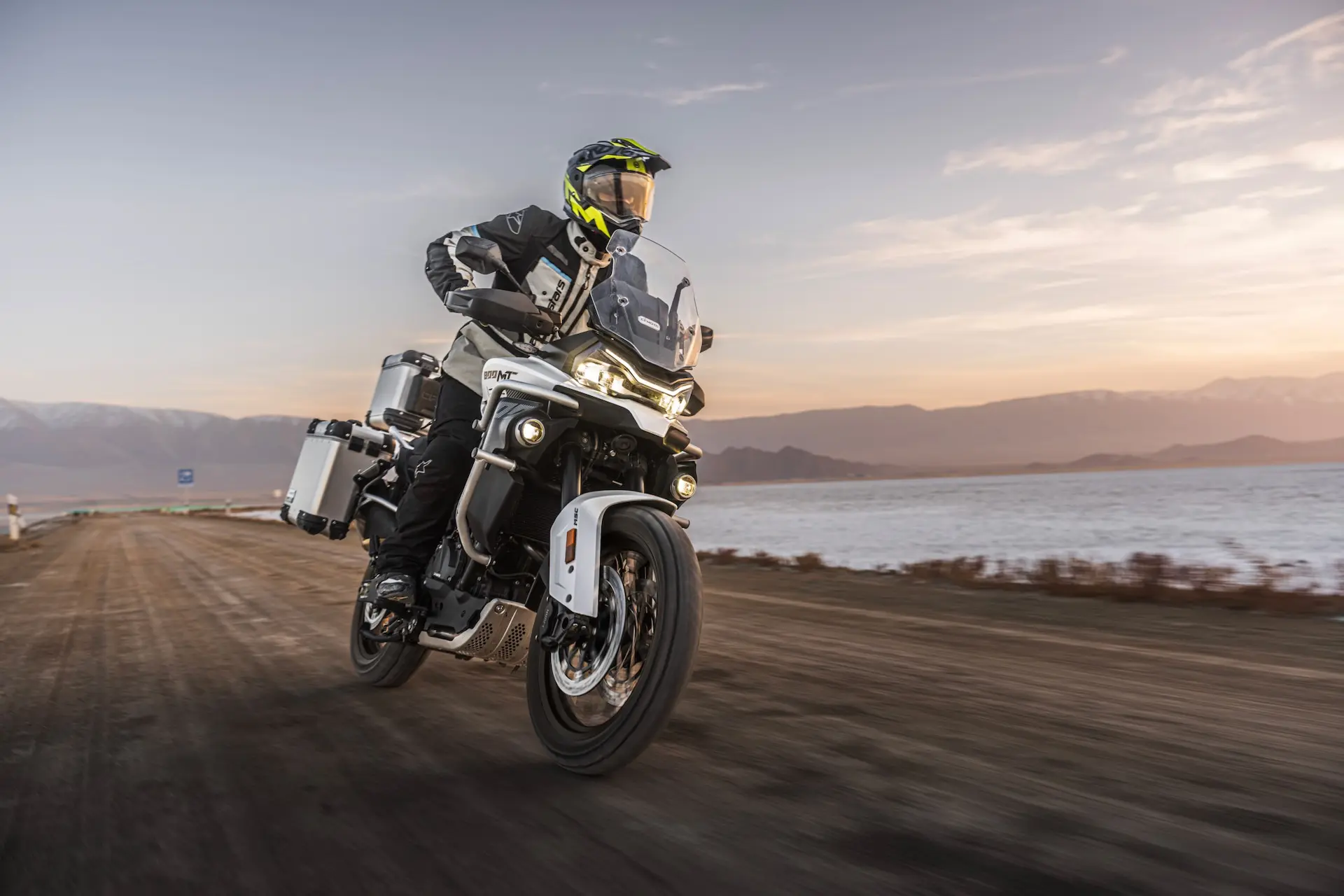 A motorcyclist wearing safety gear rides an 800MT-X adventure motorbike on a dirt track near a body of water at sunset.
