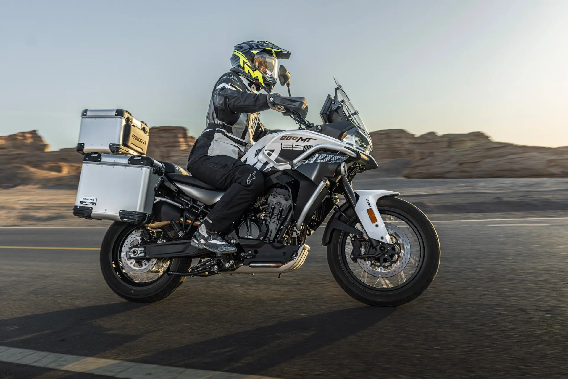 A motorcyclist wearing protective gear rides an 800MT-X adventure motorbike with panniers on a tarmacked road through a rocky desert landscape.