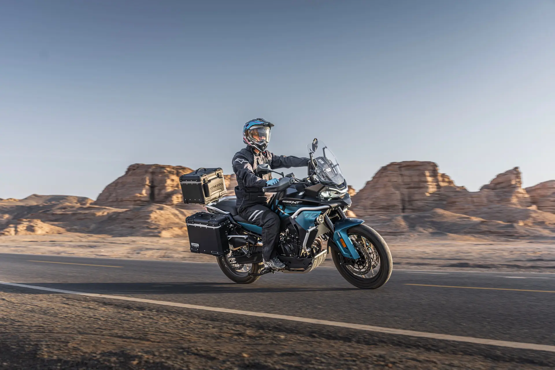 A person wearing protective gear rides an 800MT-X touring motorbike on a paved road through a desert landscape with rocky formations in the background.