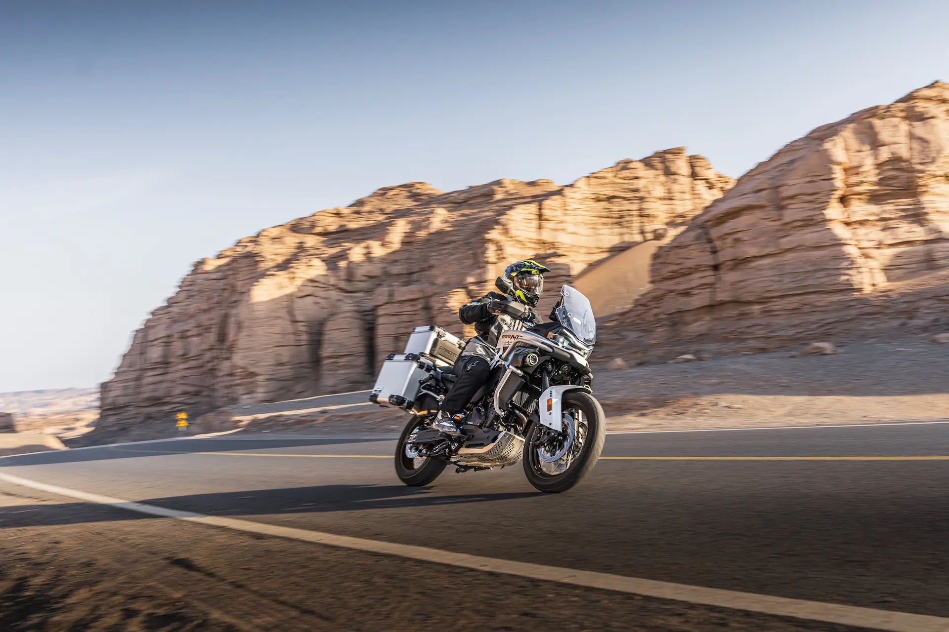 A motorcyclist wearing protective gear rides an 800MT-X touring motorbike on a tarmac road beside rocky desert cliffs under a clear sky.