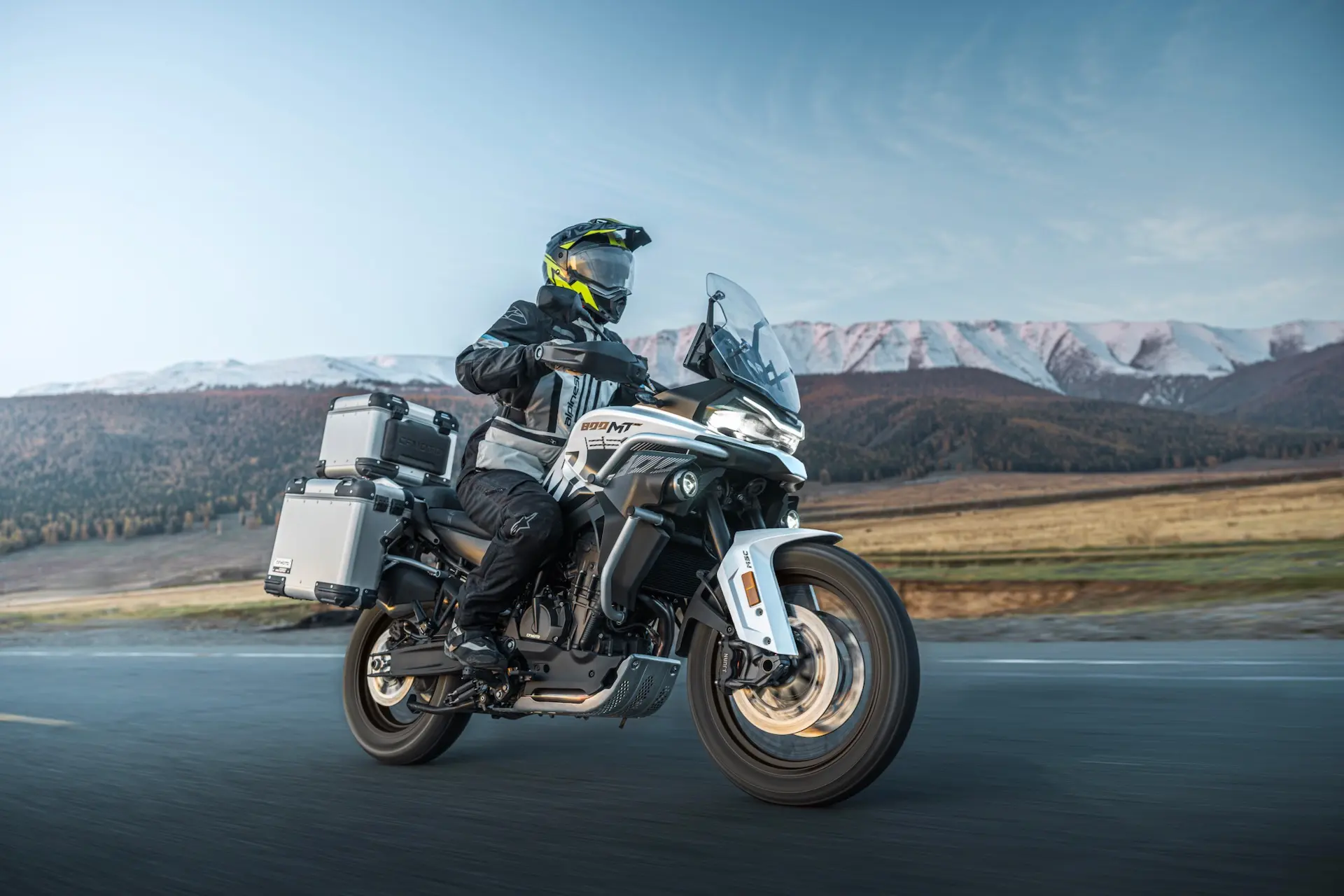 A person wearing protective gear and a helmet rides an 800MT-X touring motorbike with panniers on a road, mountains and open landscape stretching in the background.