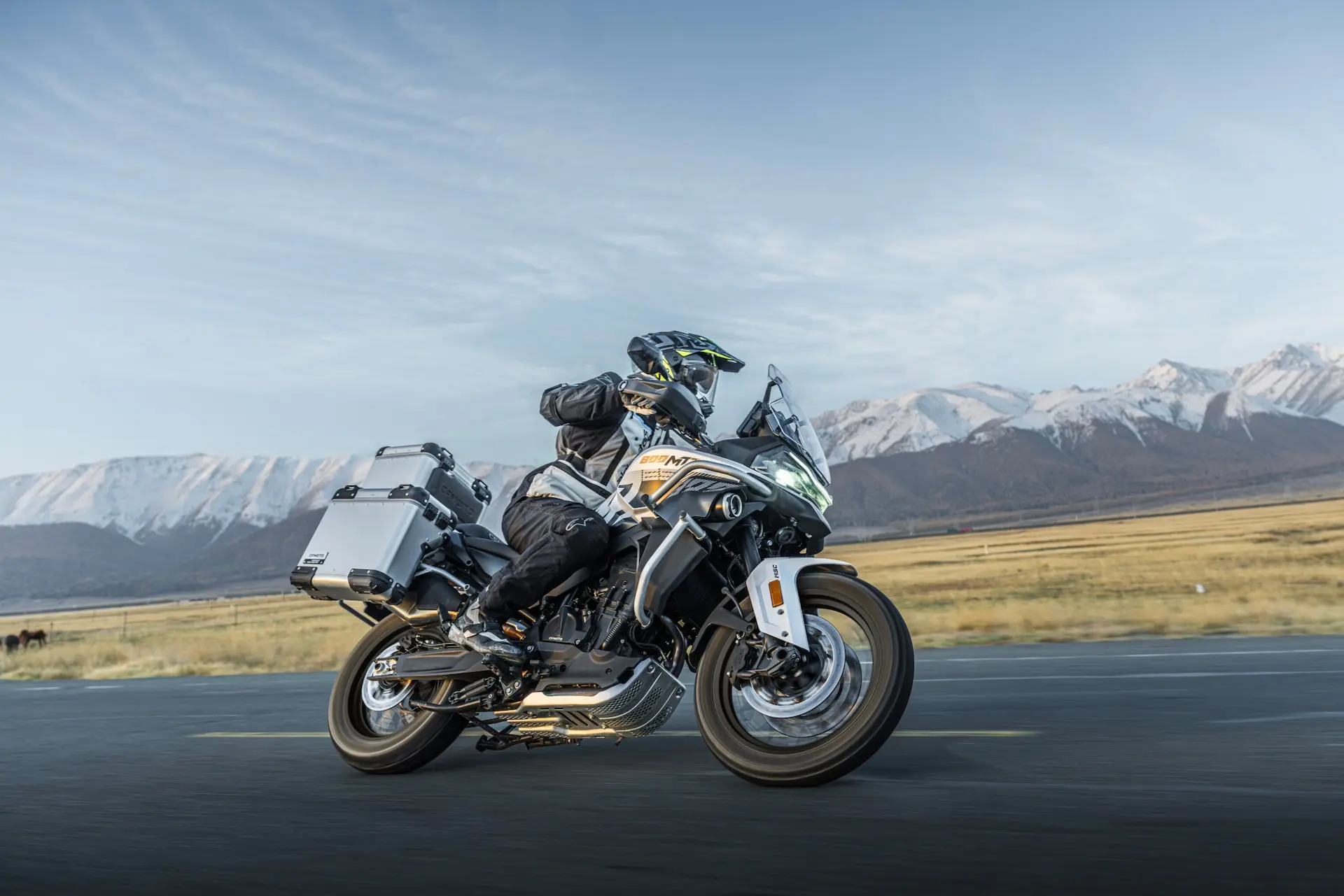 A person rides an 800MT-X touring motorbike on a tarmacked road, with snow-capped mountains and open fields in the background.