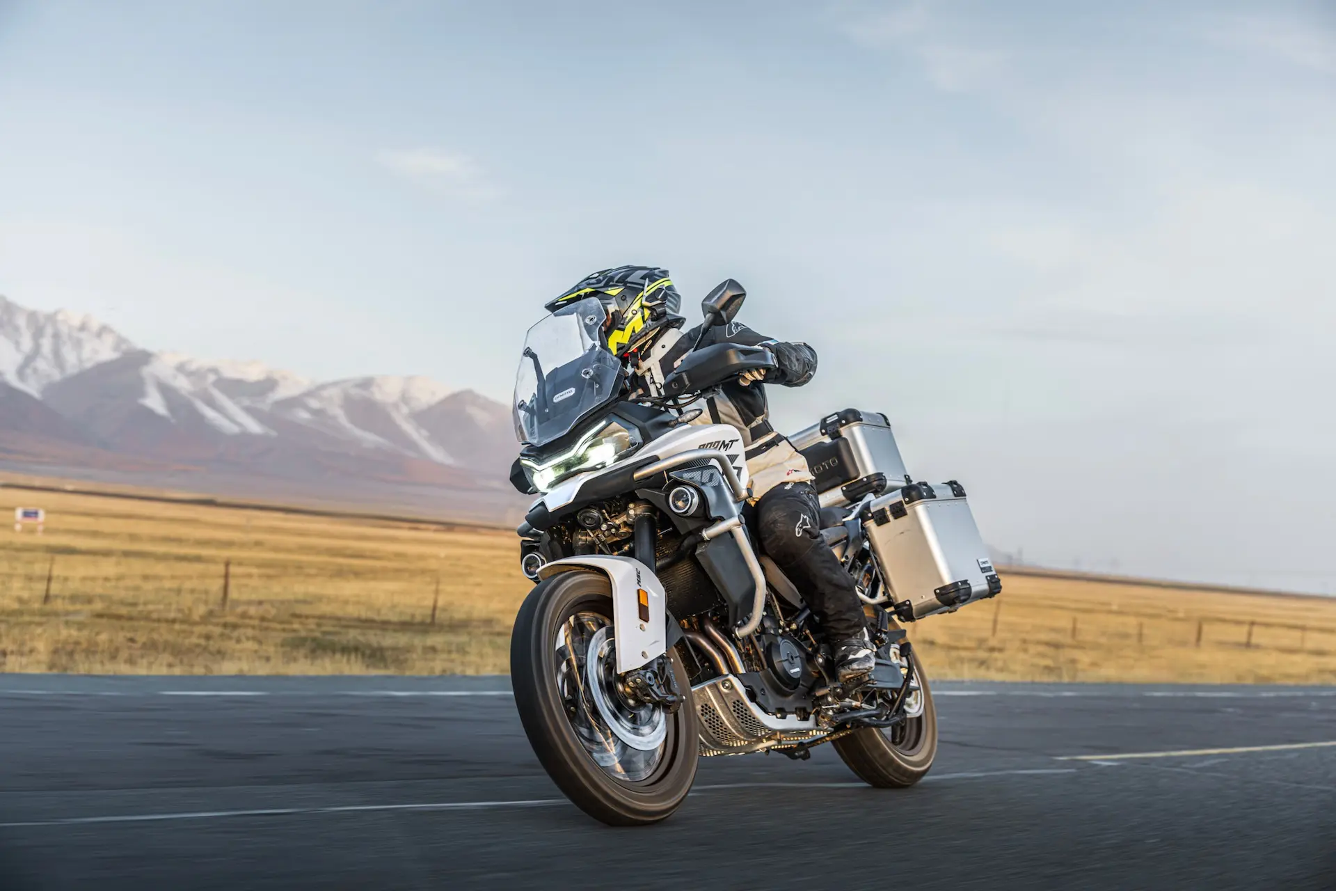 A person rides an 800MT-X touring motorbike with side and rear cargo boxes on a tarmacked road, with mountains and open fields in the background.