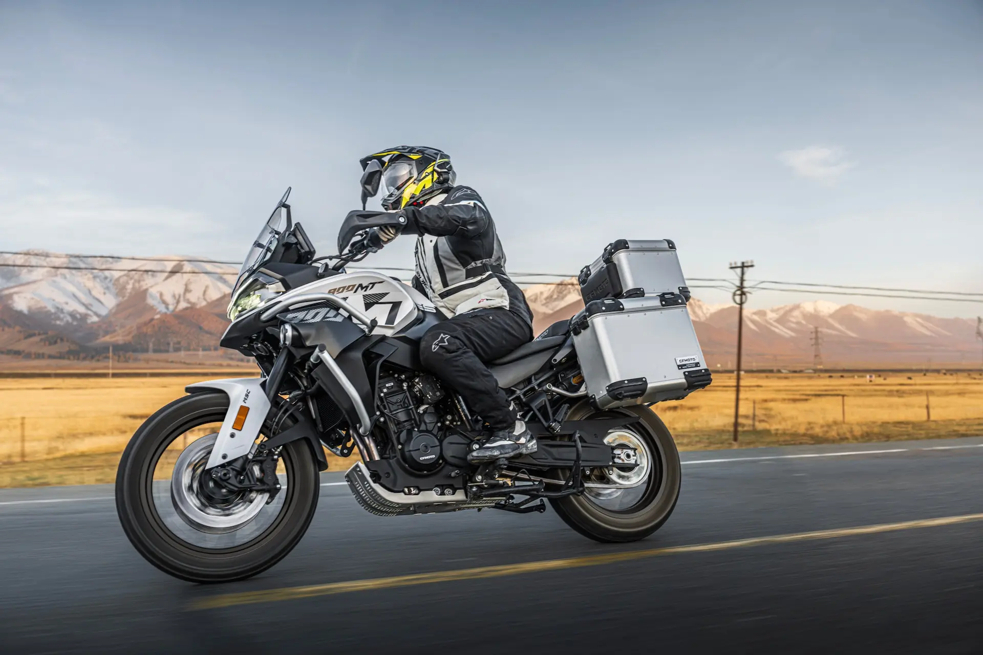 A motorcyclist wearing protective gear rides a silver 800MT-X adventure motorbike with luggage cases on an open road, with mountains and fields in the background.